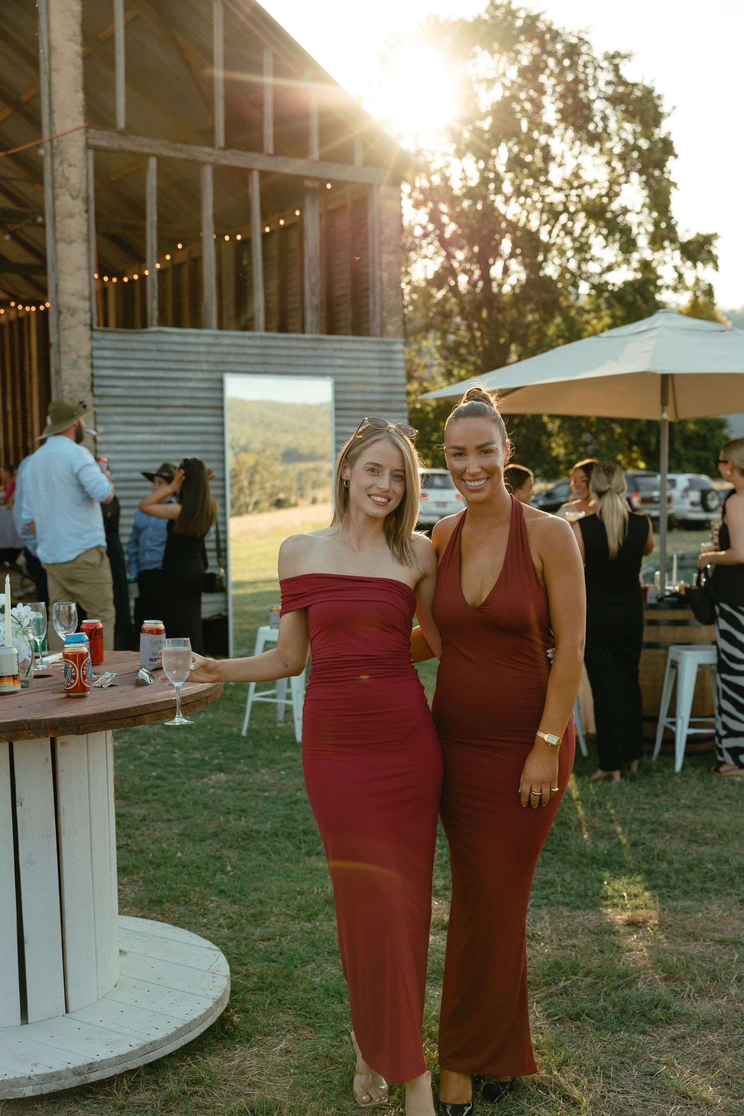 Two women in elegant red dresses pose at an outdoor event with string lights and sunset in the background at sunset.