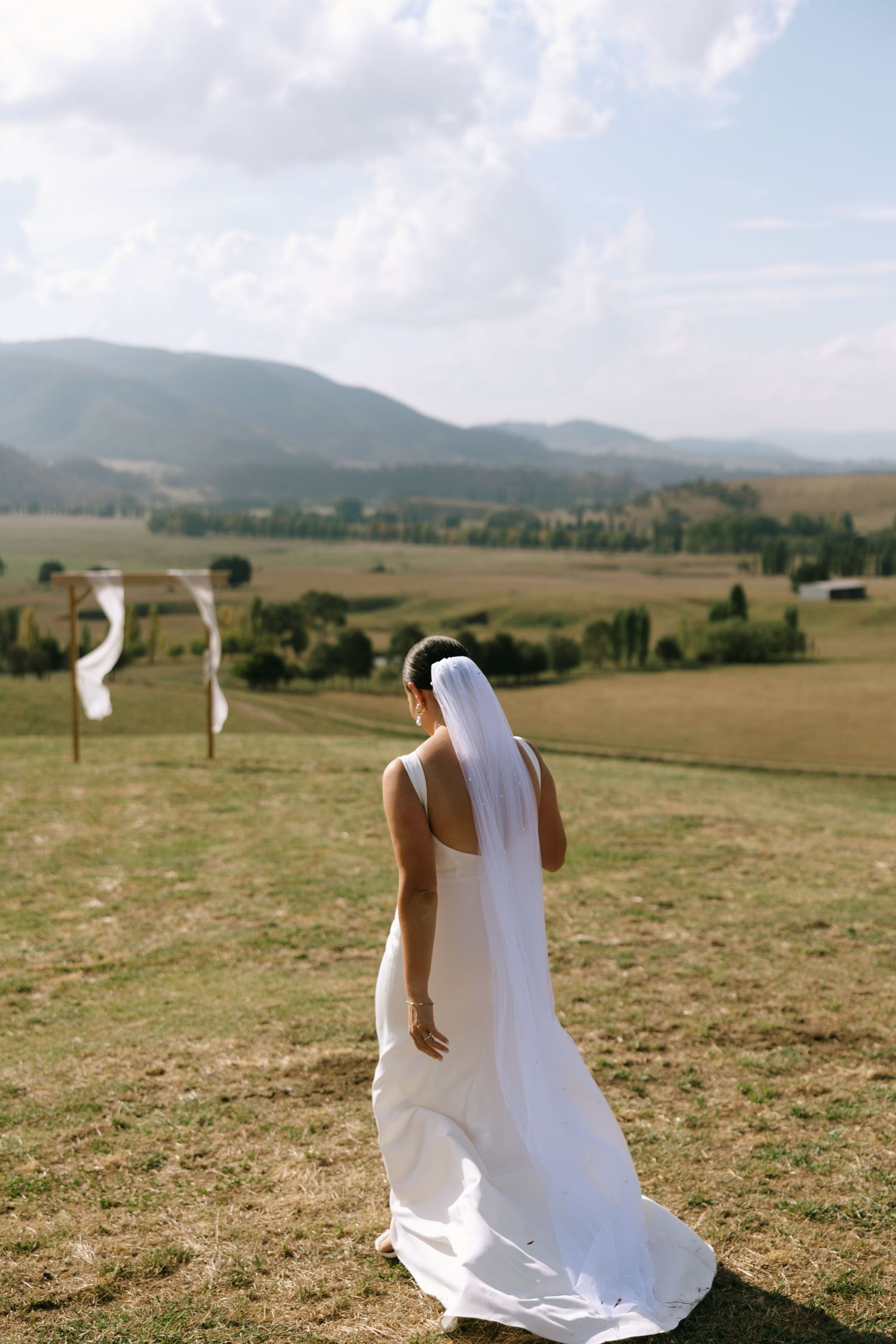 Bride in flowing white dress looking towards mountain vista and wooden ceremony arch.