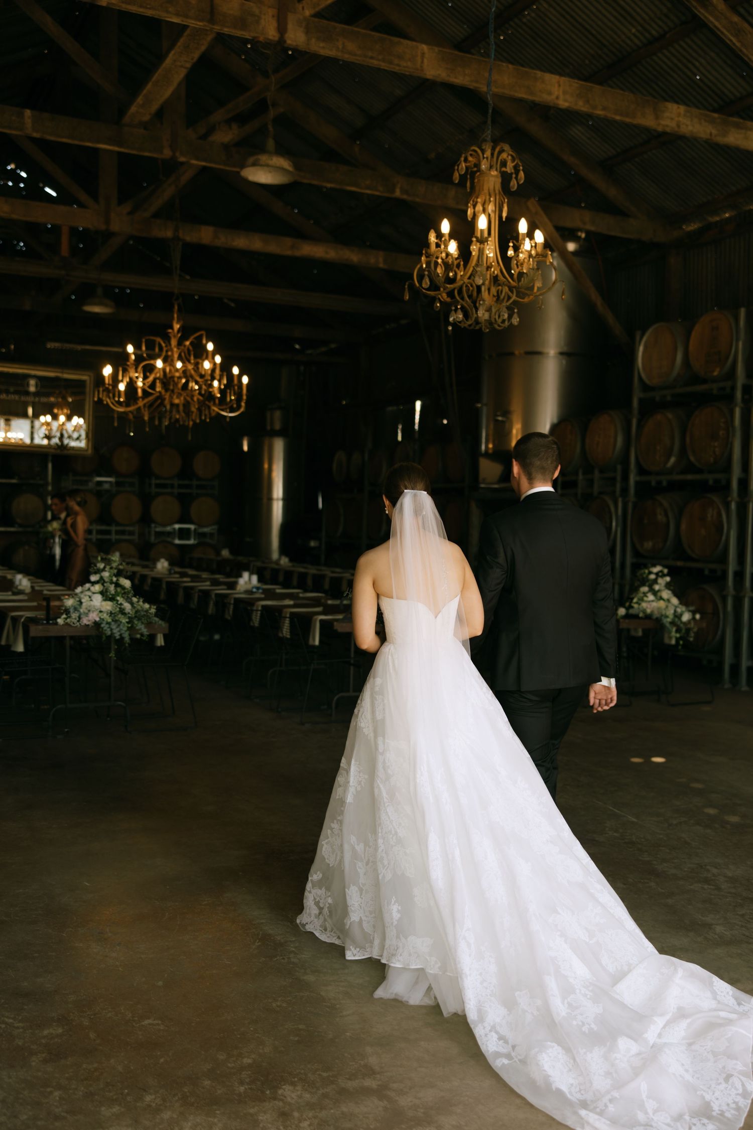 A couple walks through a rustic winery venue with elegant chandeliers and wine barrels.