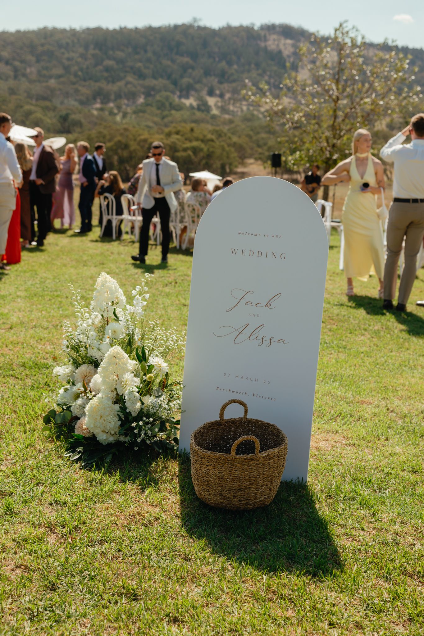 An elegant wedding welcome sign with white flowers and wicker basket displayed on grassy lawn with guests mingling in background.
