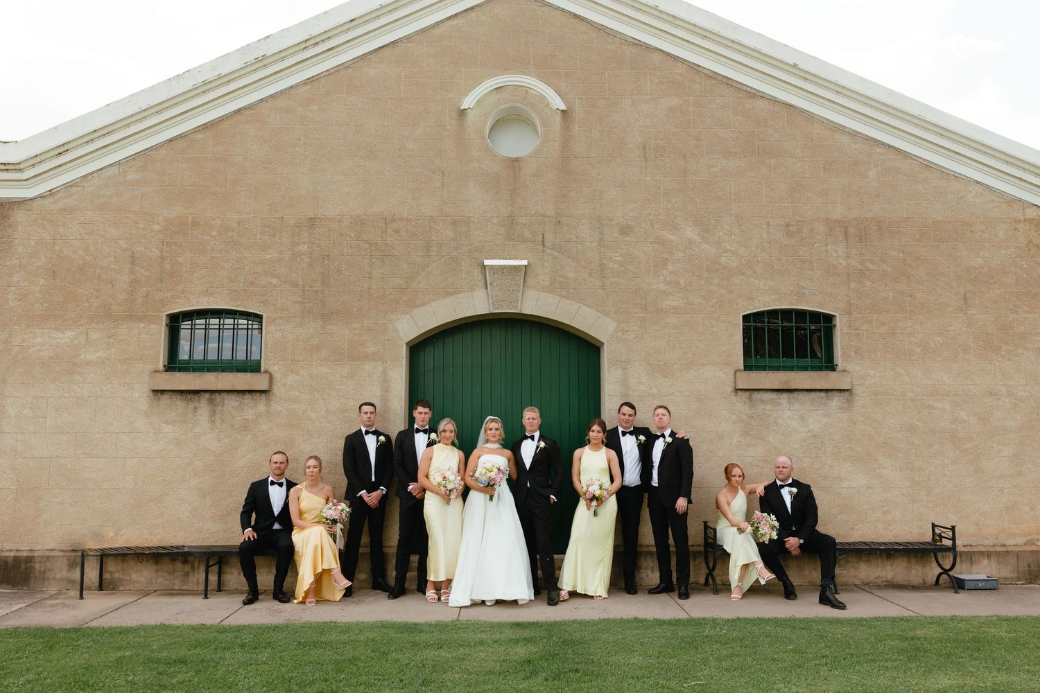 Wedding party stands in front of a beige building with a green door and small windows at a rustic venue.