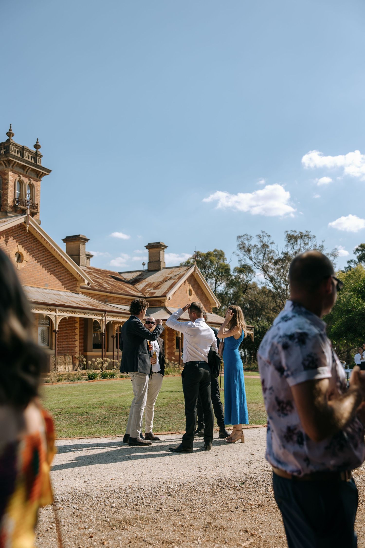 Wedding guests gather outside a historic sandstone mansion on a sunny day with blue skies.
