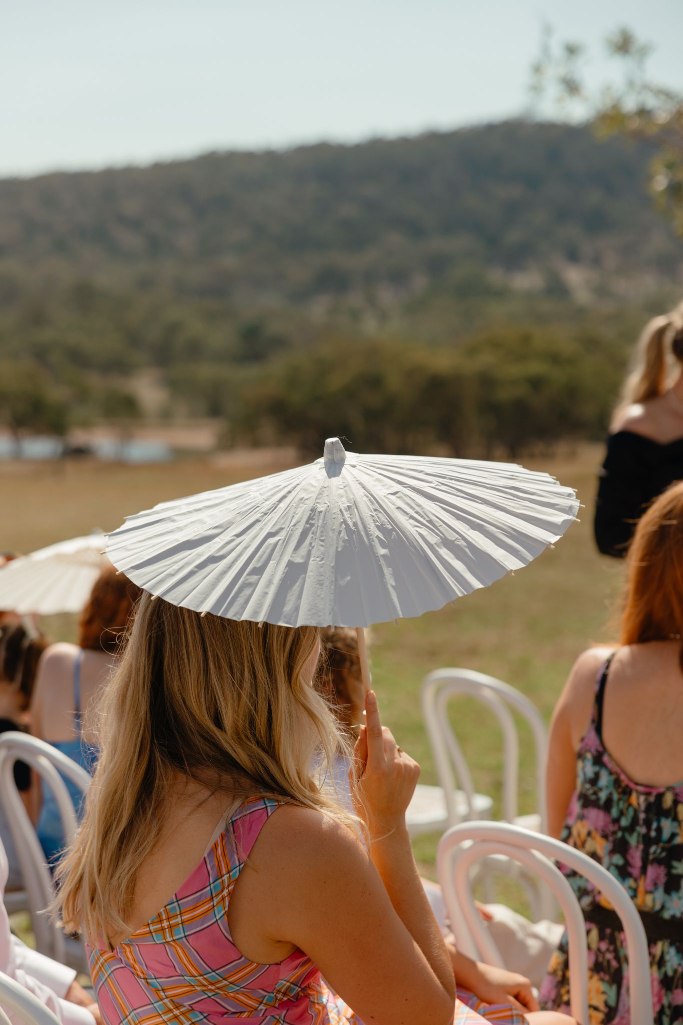 Wedding guest shielding from sun with white paper parasol while seated outdoors against mountain backdrop.