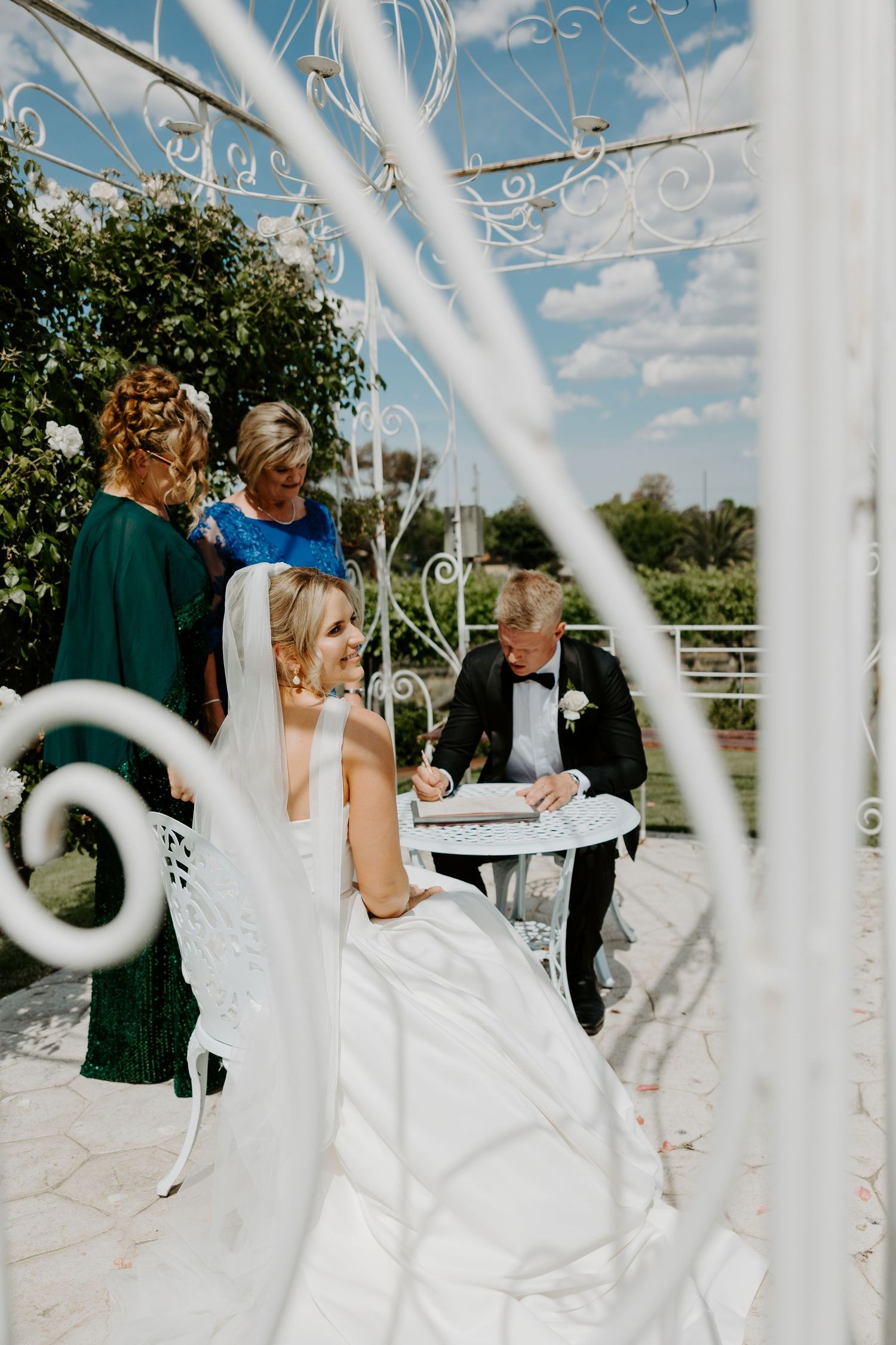 Wedding ceremony taking place in an ornate white gazebo with decorative ironwork details under blue sky.