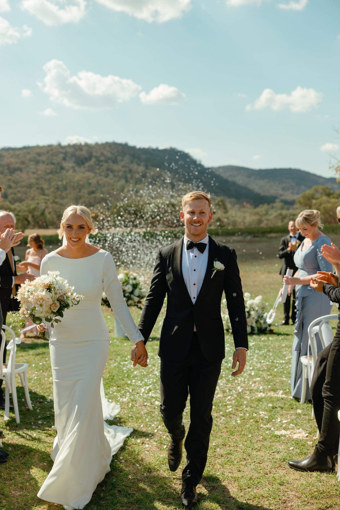 A newlywed couple exits their outdoor mountain ceremony while guests throw flower petals.