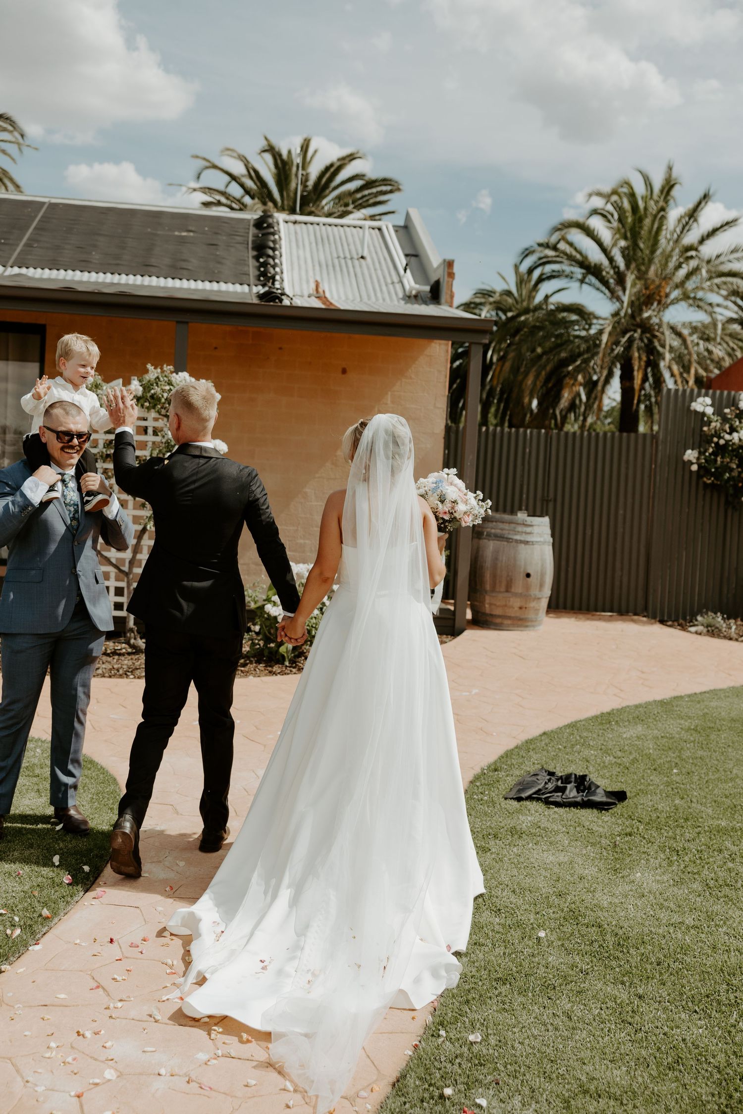 Outdoor wedding ceremony in a garden setting with palm trees and a beautiful long white veil trailing behind the bride.