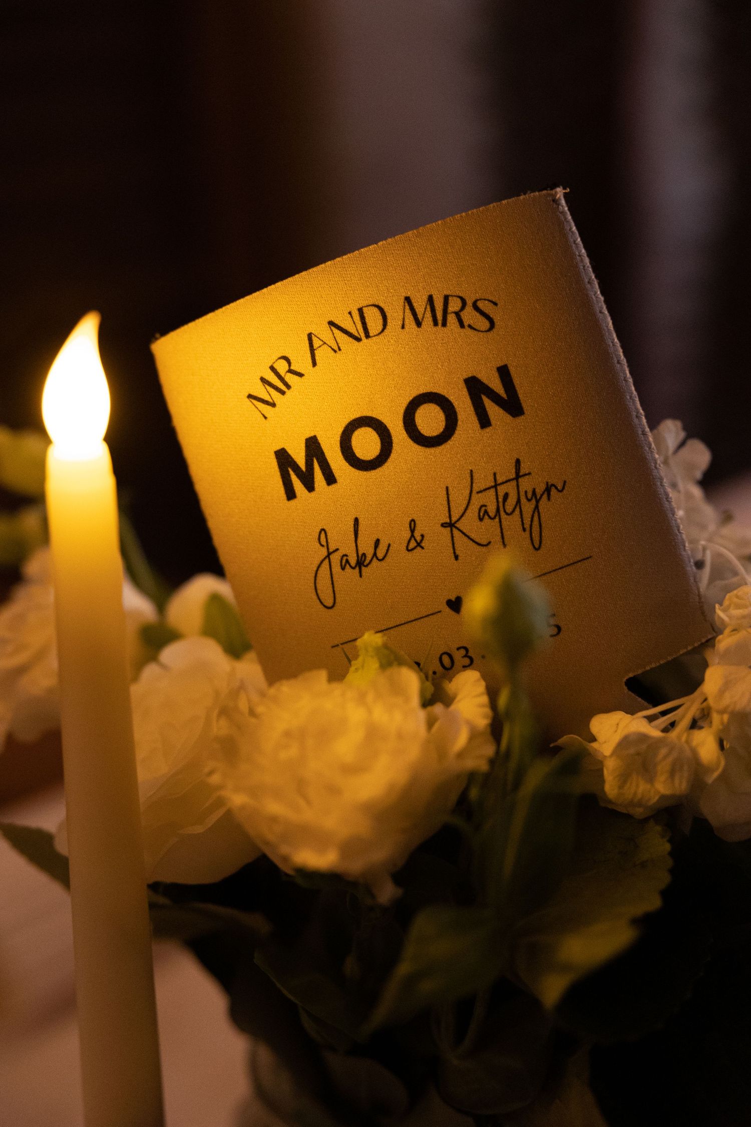 A wedding reception table setting with a lit candle and name card reading 'Mr and Mrs Moon' beside white roses.