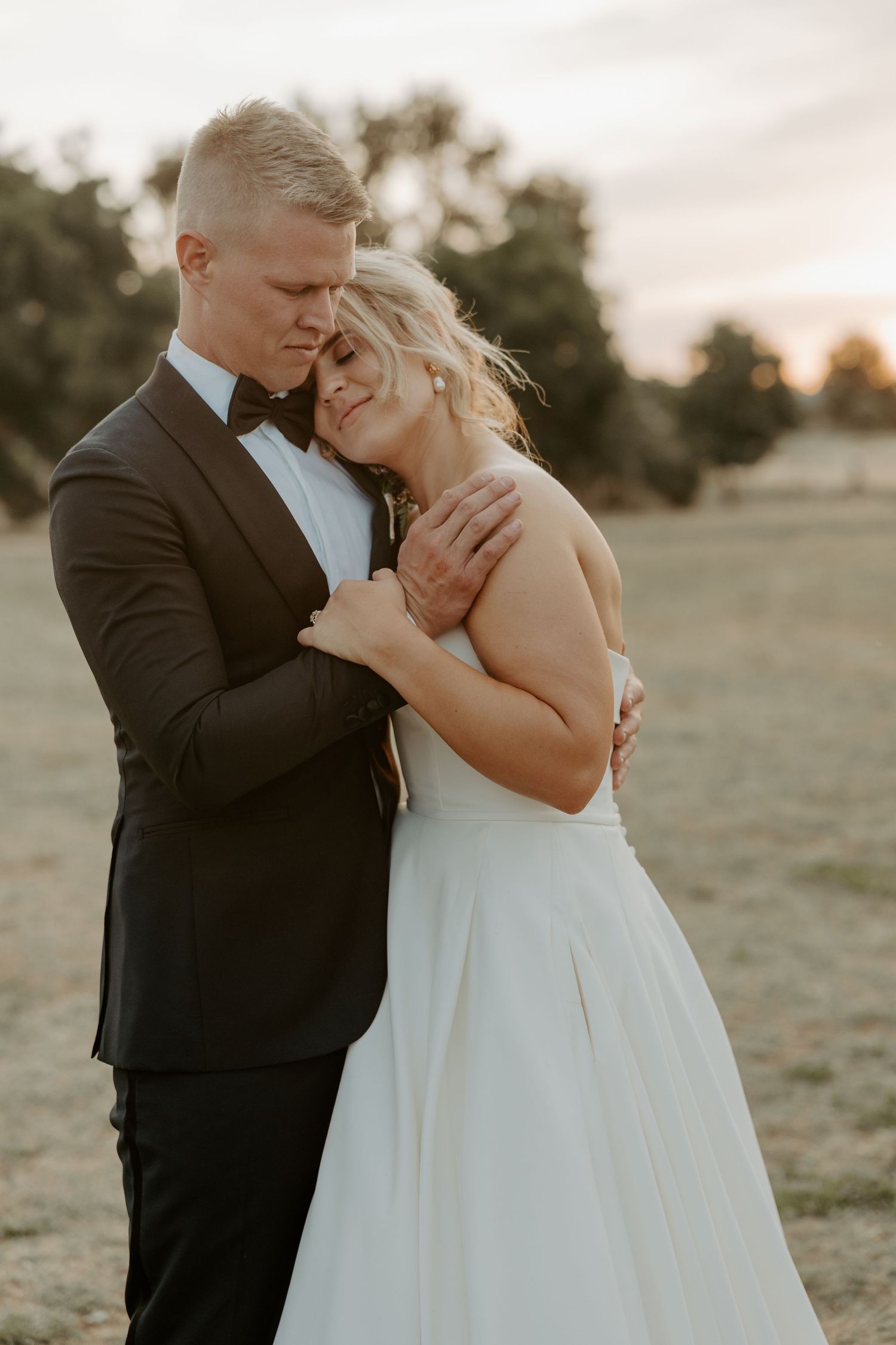 A romantic couple embraces during golden hour sunset, with the bride in white and groom in black tuxedo.
