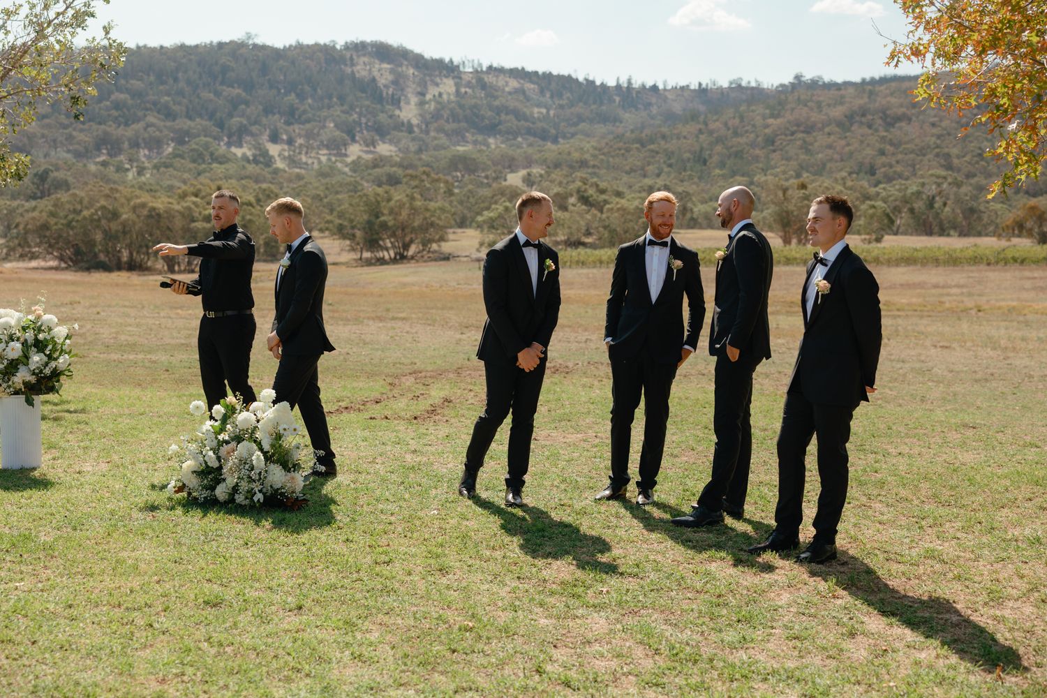 Groomsmen in black suits stand together on a grassy field with autumn mountains in the background during an outdoor wedding.