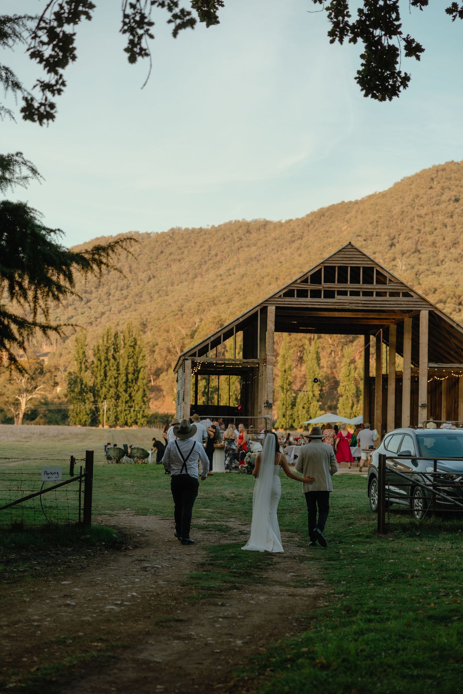 Wedding guests walk toward a rustic barn venue with mountains and golden sunset light in the background.