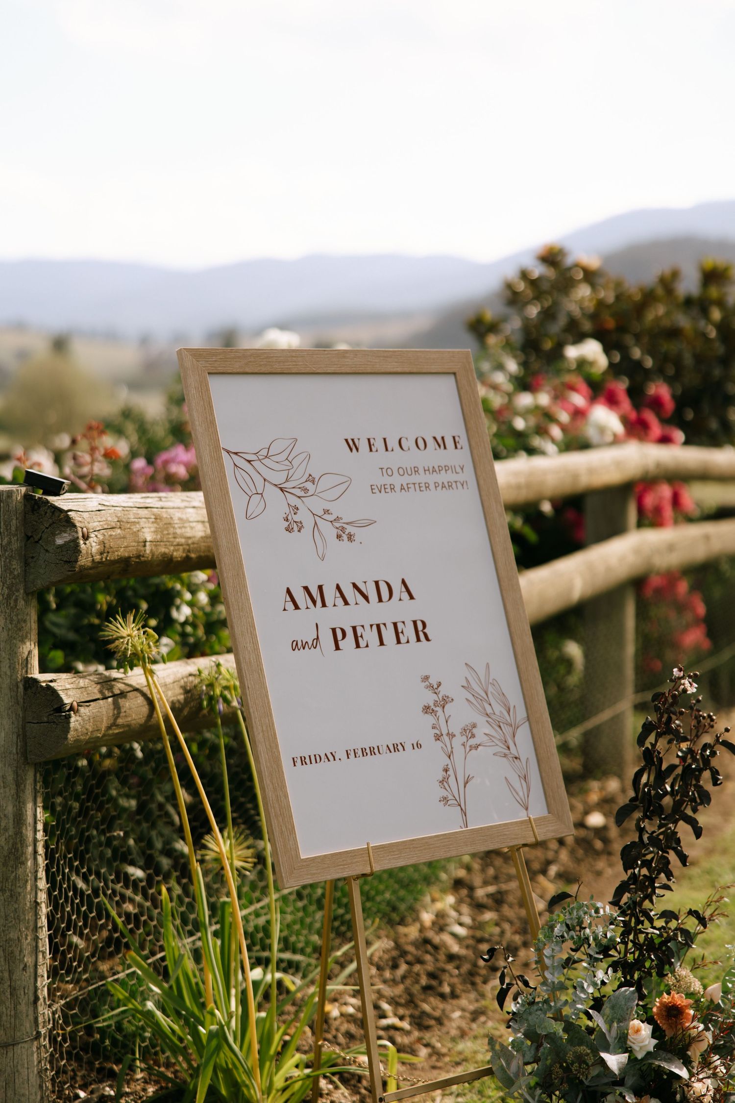 A rustic wedding welcome sign with floral details displayed on a wooden fence with mountains in the background.