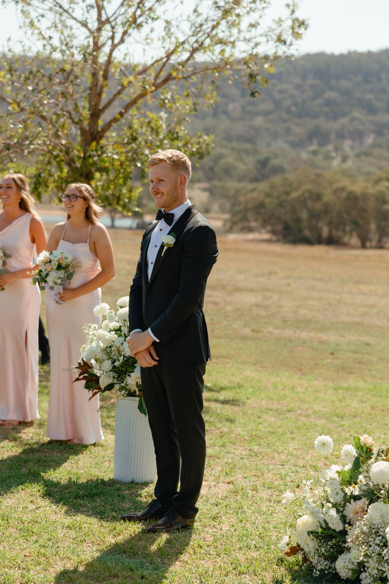 Wedding ceremony outdoors with bridesmaids in pink dresses and white floral arrangements in natural landscape.
