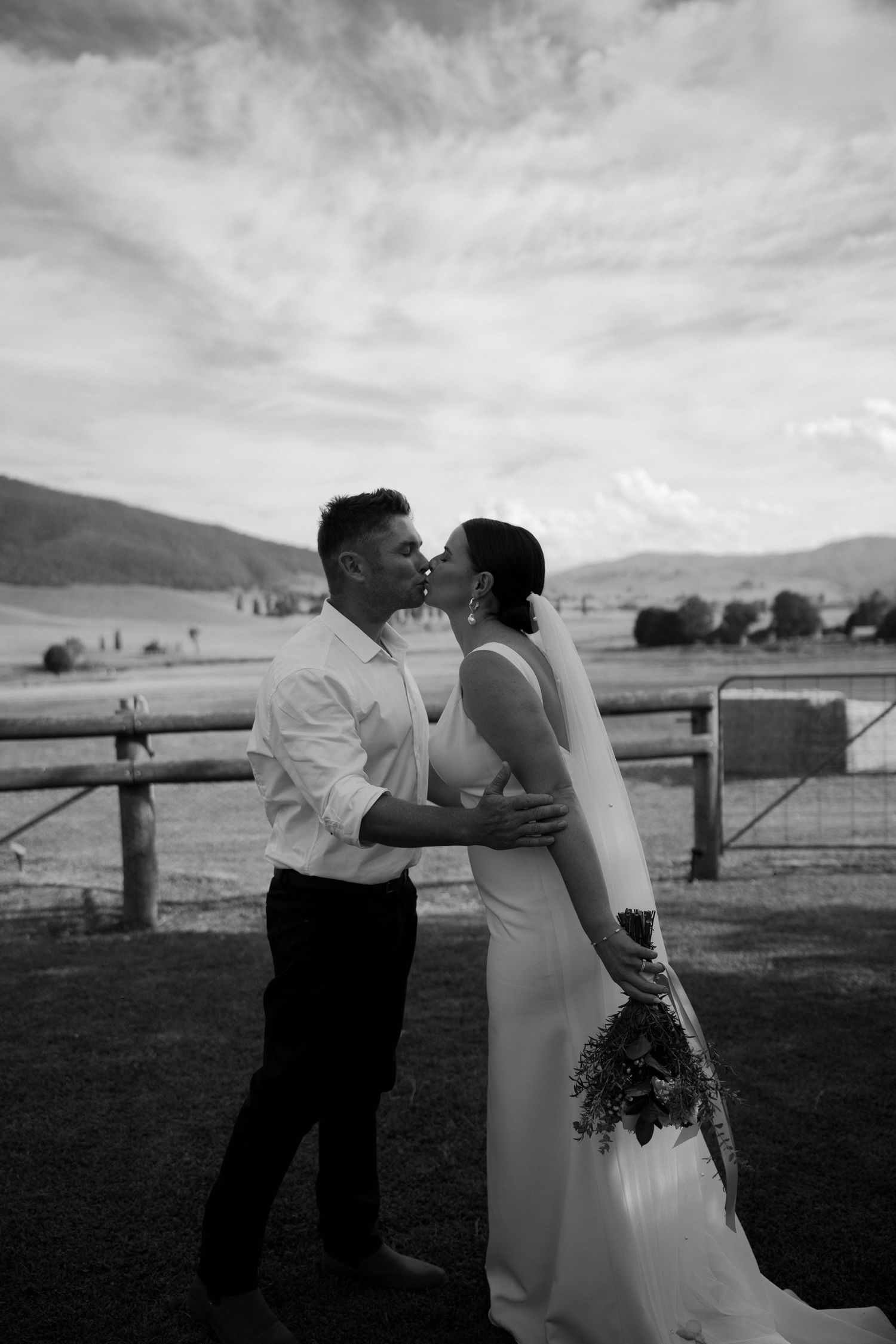 A romantic black and white wedding photo of a couple embracing on a ranch with mountains in the background.