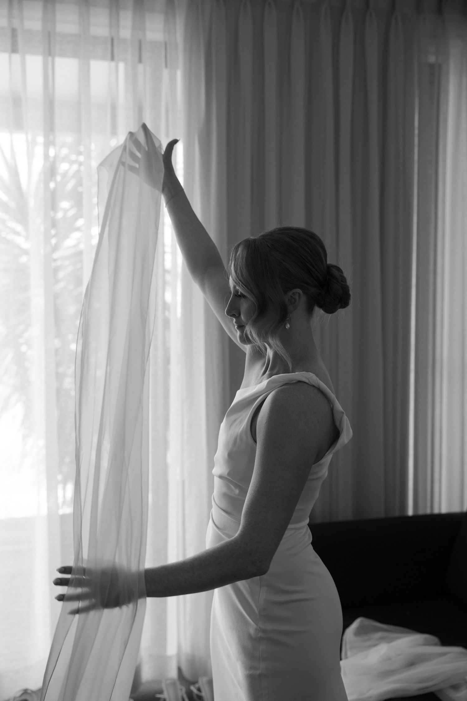 A person in an elegant white dress reaches up to touch sheer curtains in a bright room with soft lighting, captured in black and white.
