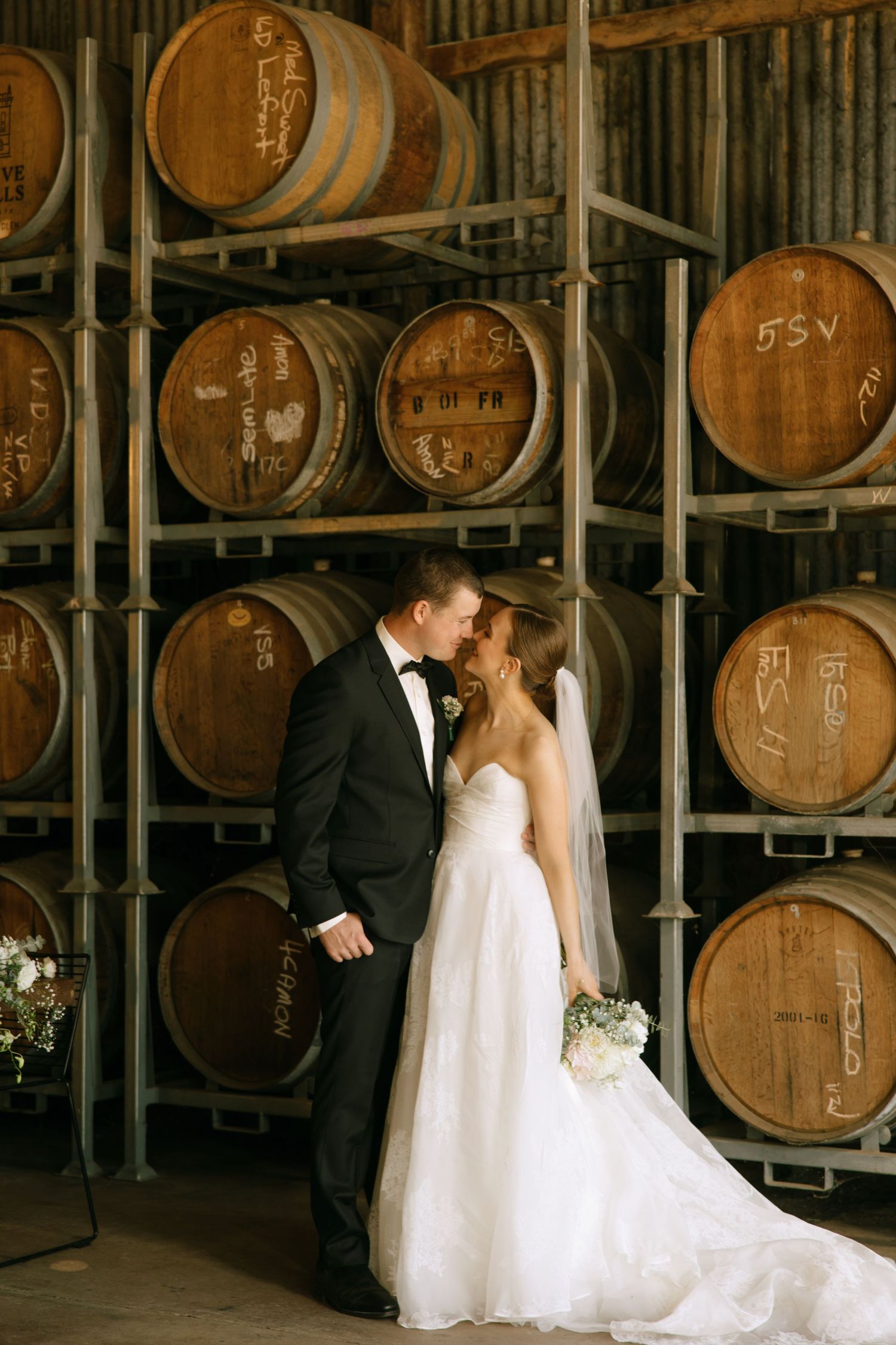 A newlywed couple shares an intimate moment in front of stacked wine barrels at a winery venue.