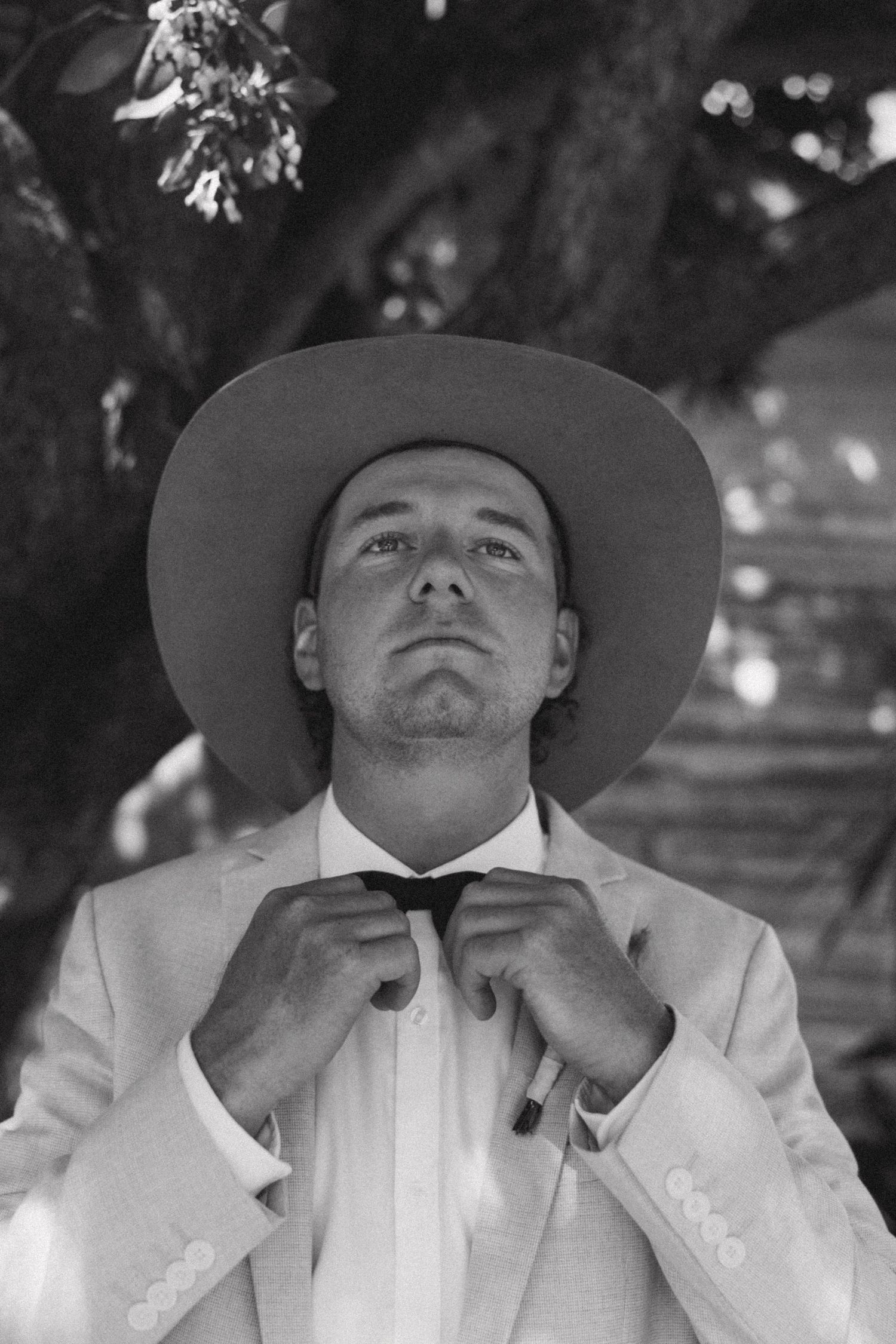 Classic black and white portrait of a groom adjusting his bowtie in a cowboy hat.