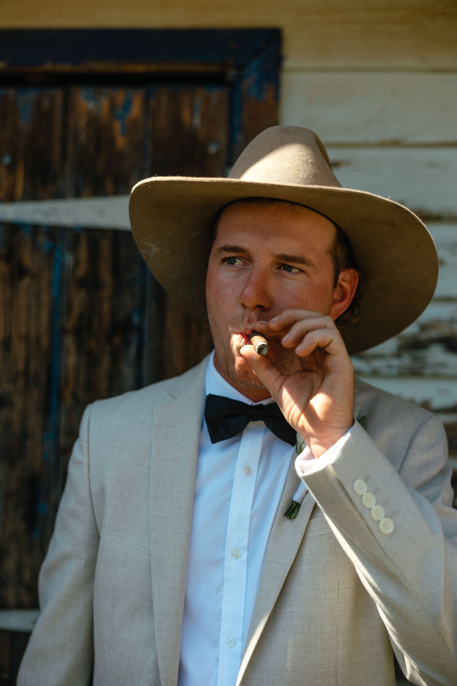 Groom in a light colored suit and cowboy hat smoking outdoors.