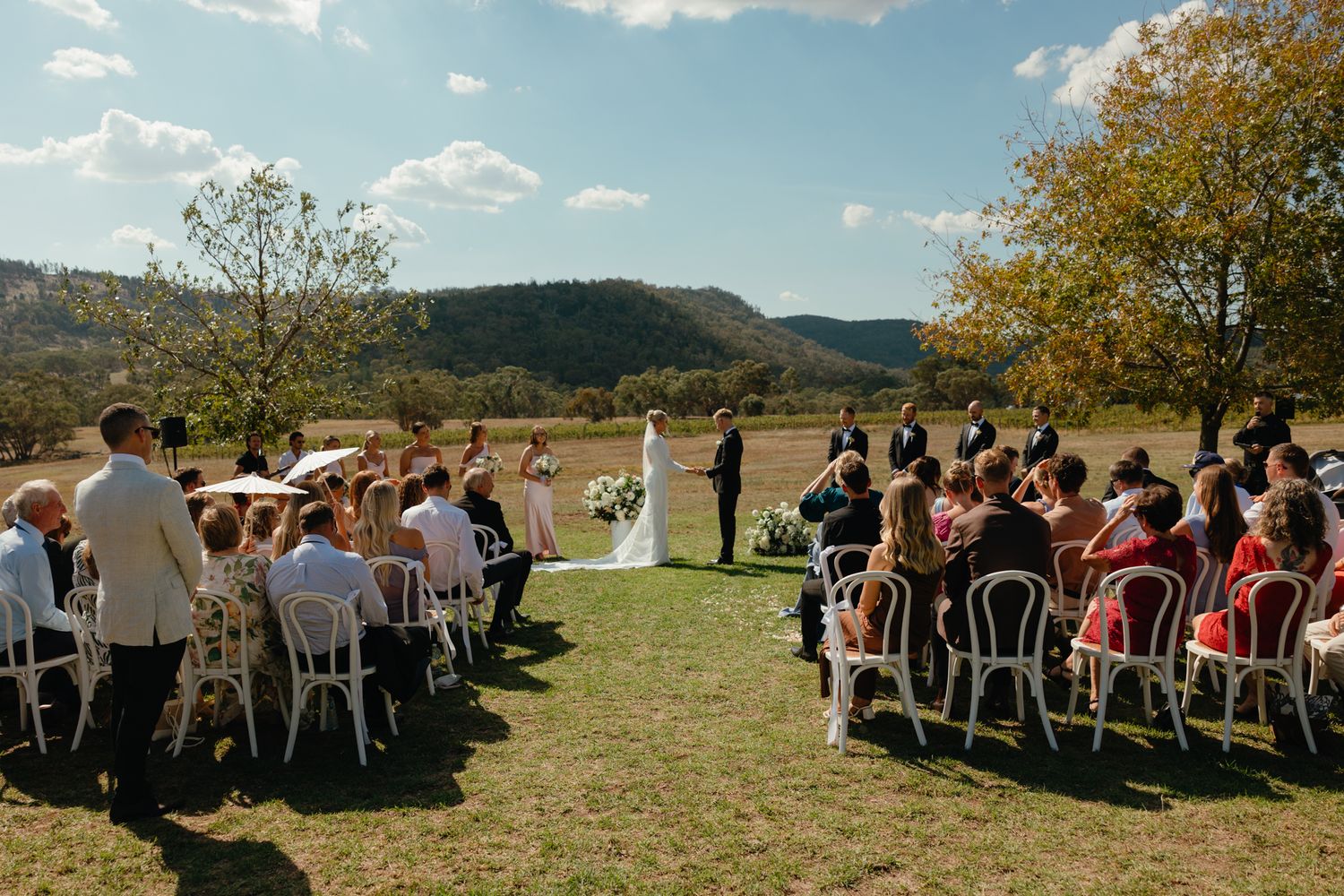Outdoor wedding ceremony in progress with guests seated on white chairs overlooking scenic mountain landscape.