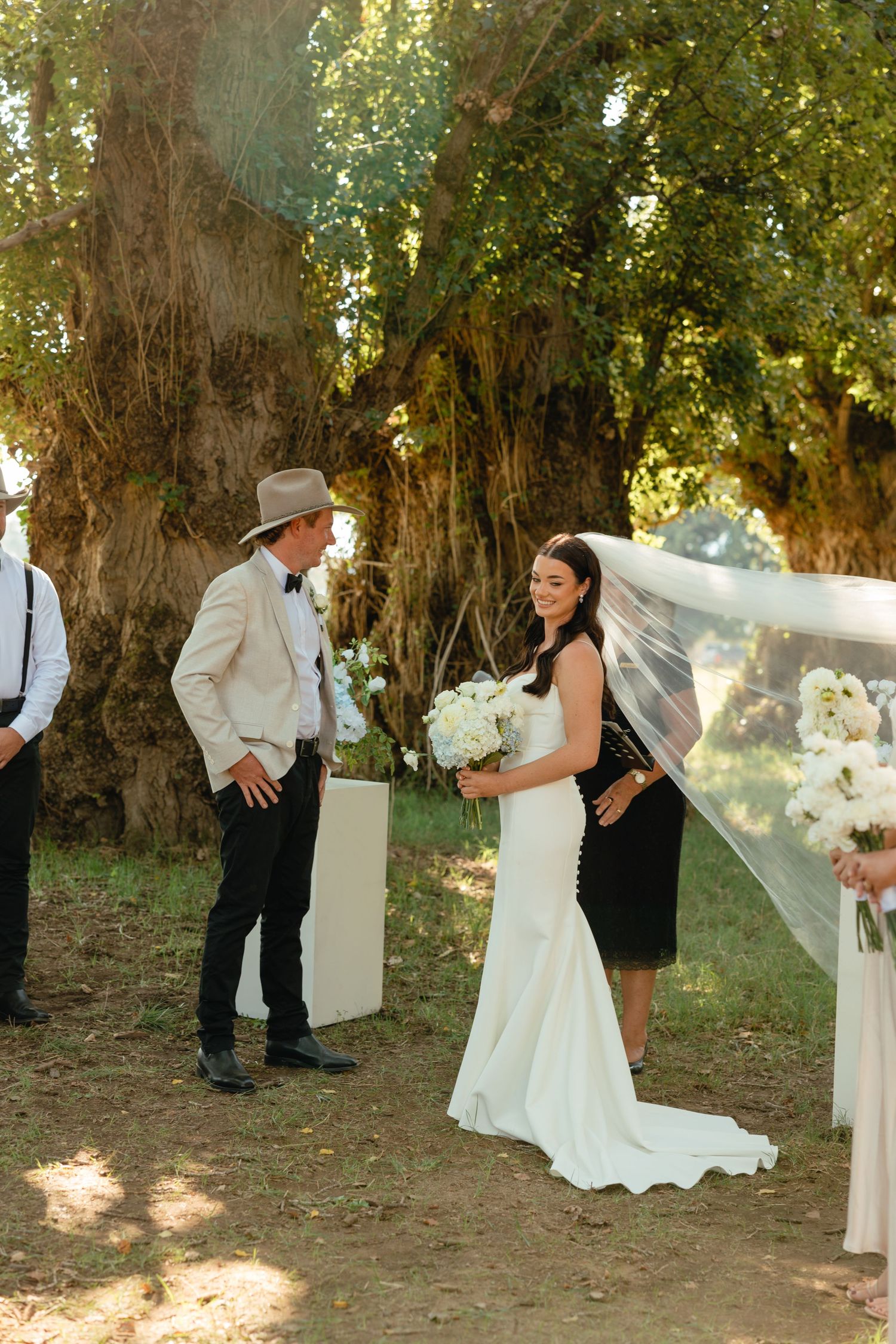 An outdoor wedding ceremony takes place among tall trees with a flowing white veil and elegant floral arrangements.