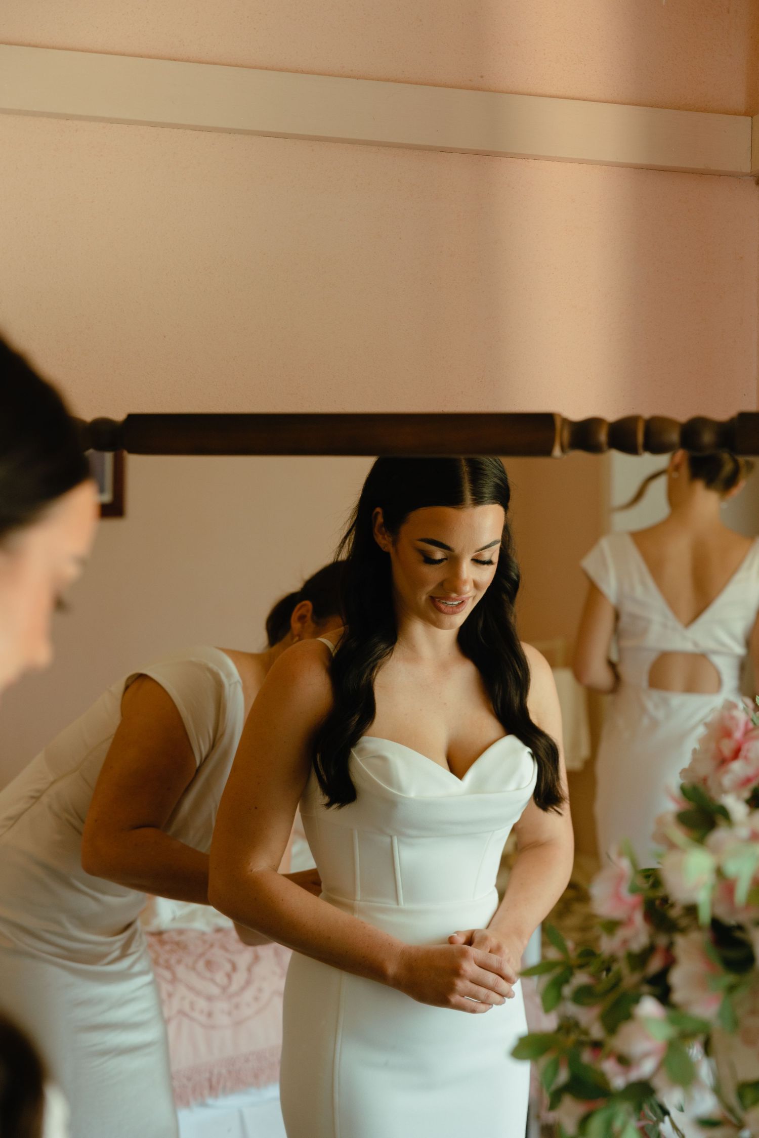 A bride in a form-fitting white dress smiles while getting ready in a room with floral decorations.
