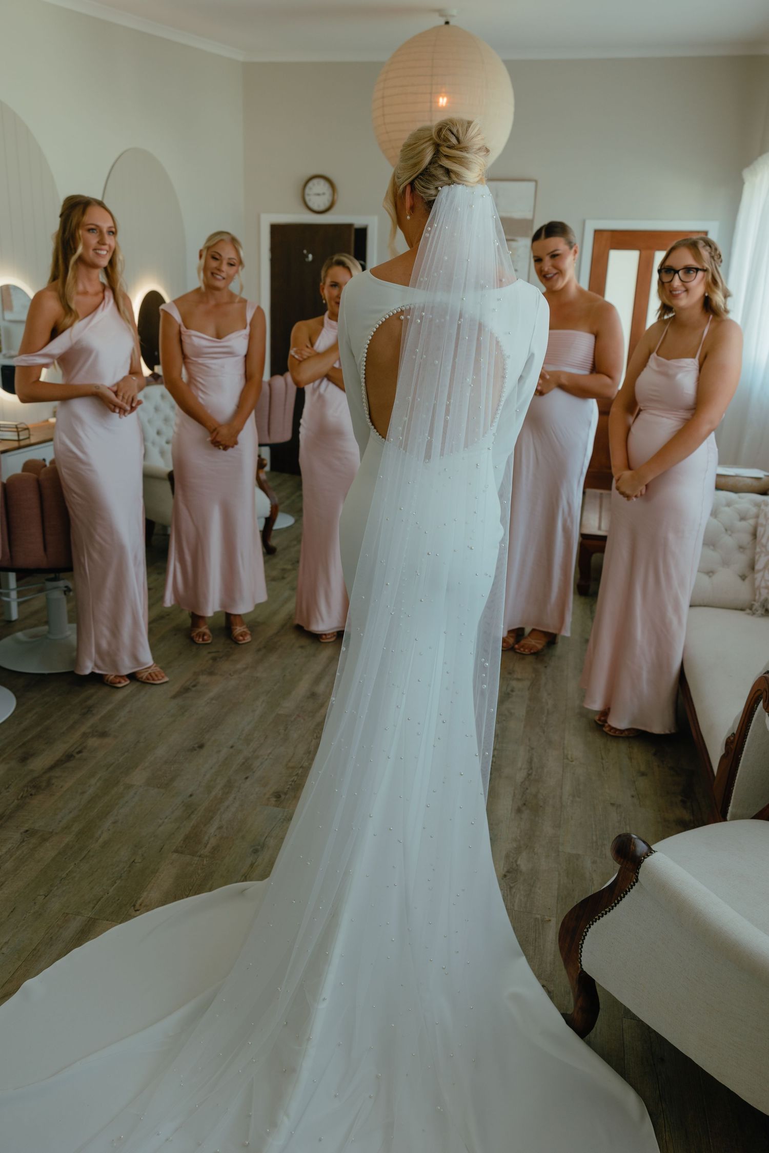 Bridesmaids in blush pink dresses admiring the bride in her flowing white wedding gown with train.