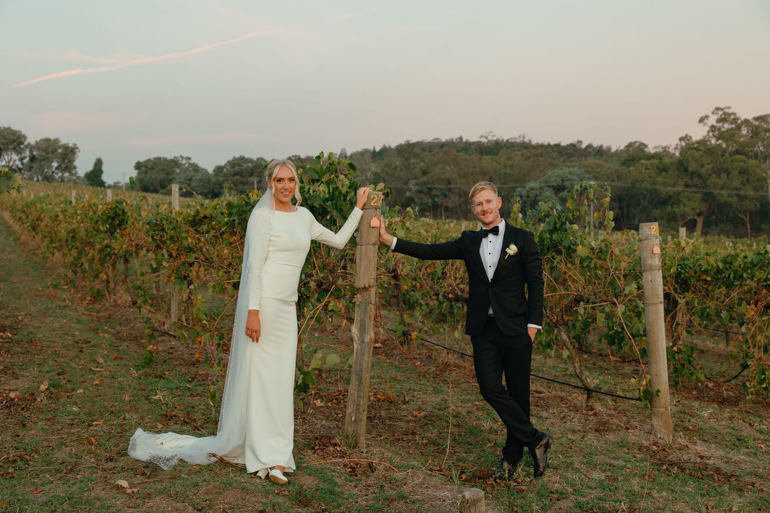 A couple stands at a rustic wooden post and wire fence overlooking a scenic vineyard at sunset.