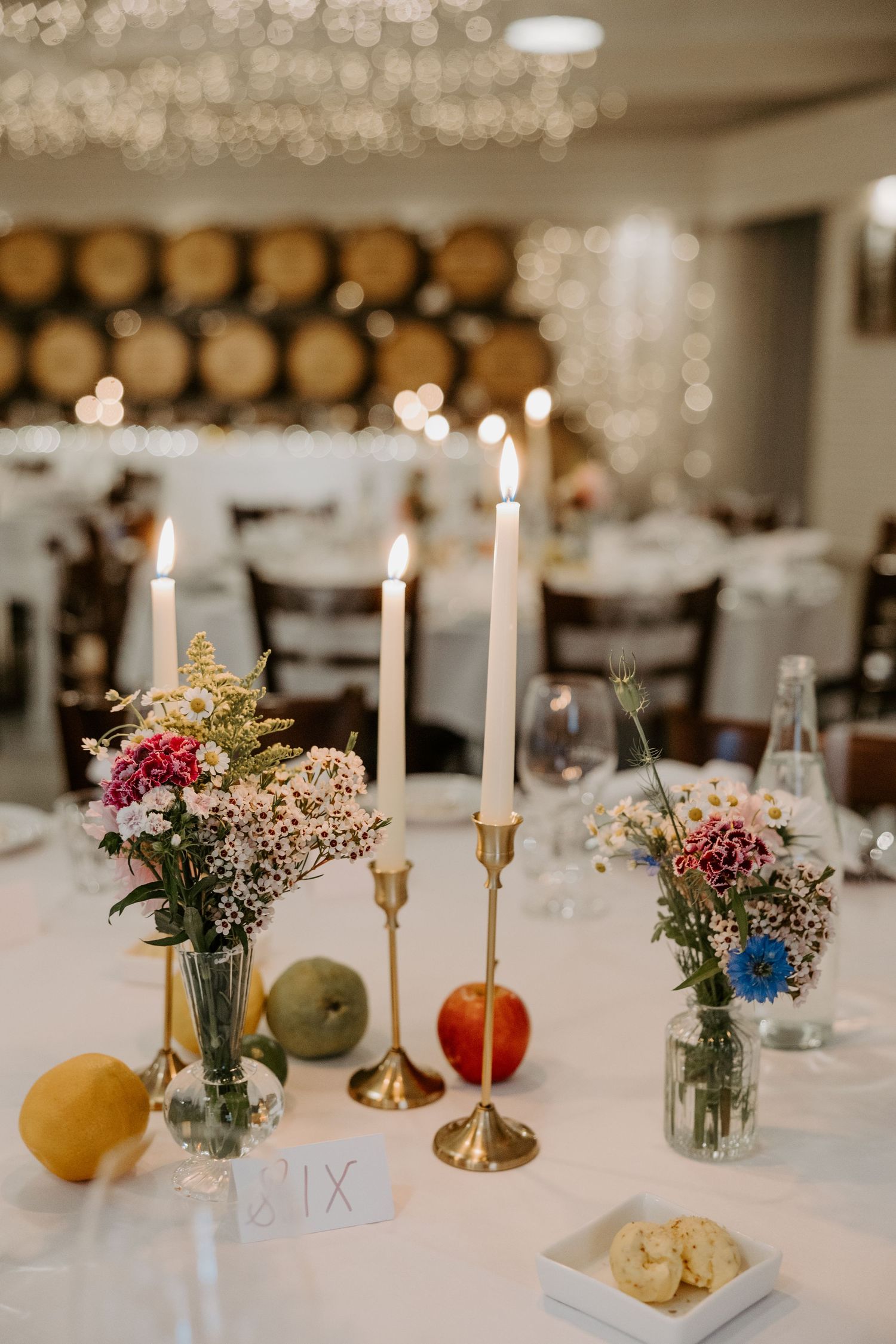 Elegant wedding reception table with white candles, small floral arrangements in mason jars, and wooden barrels backdrop.