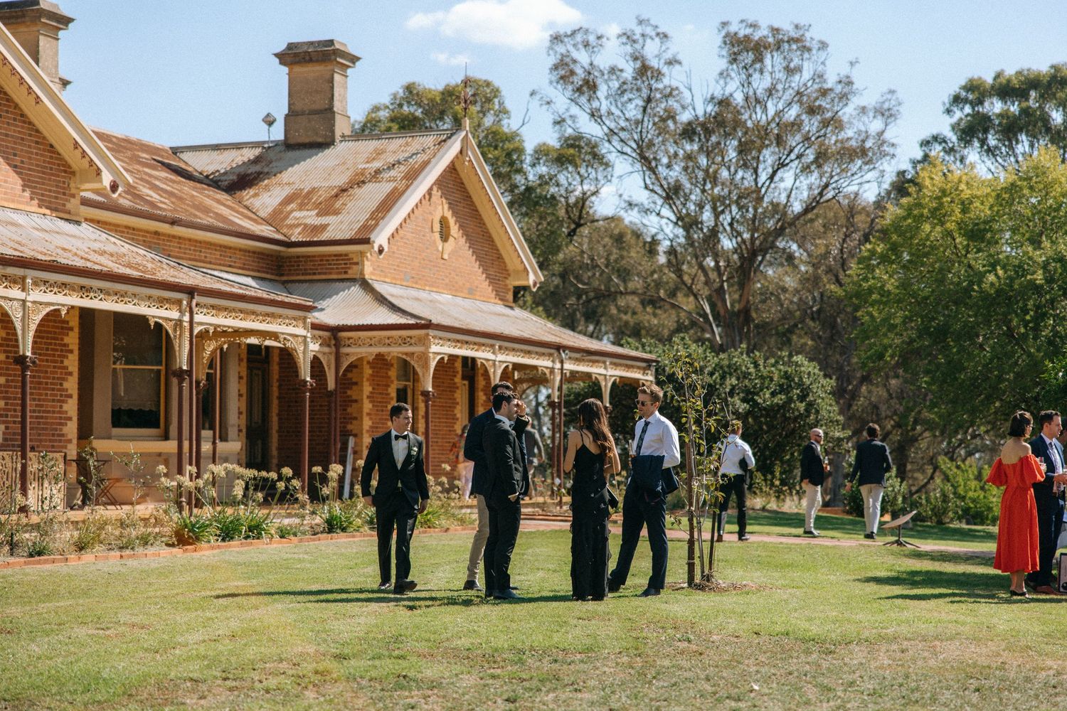 Group of guests gather on the lawn outside a historic Victorian-style mansion with wraparound veranda on a sunny day.