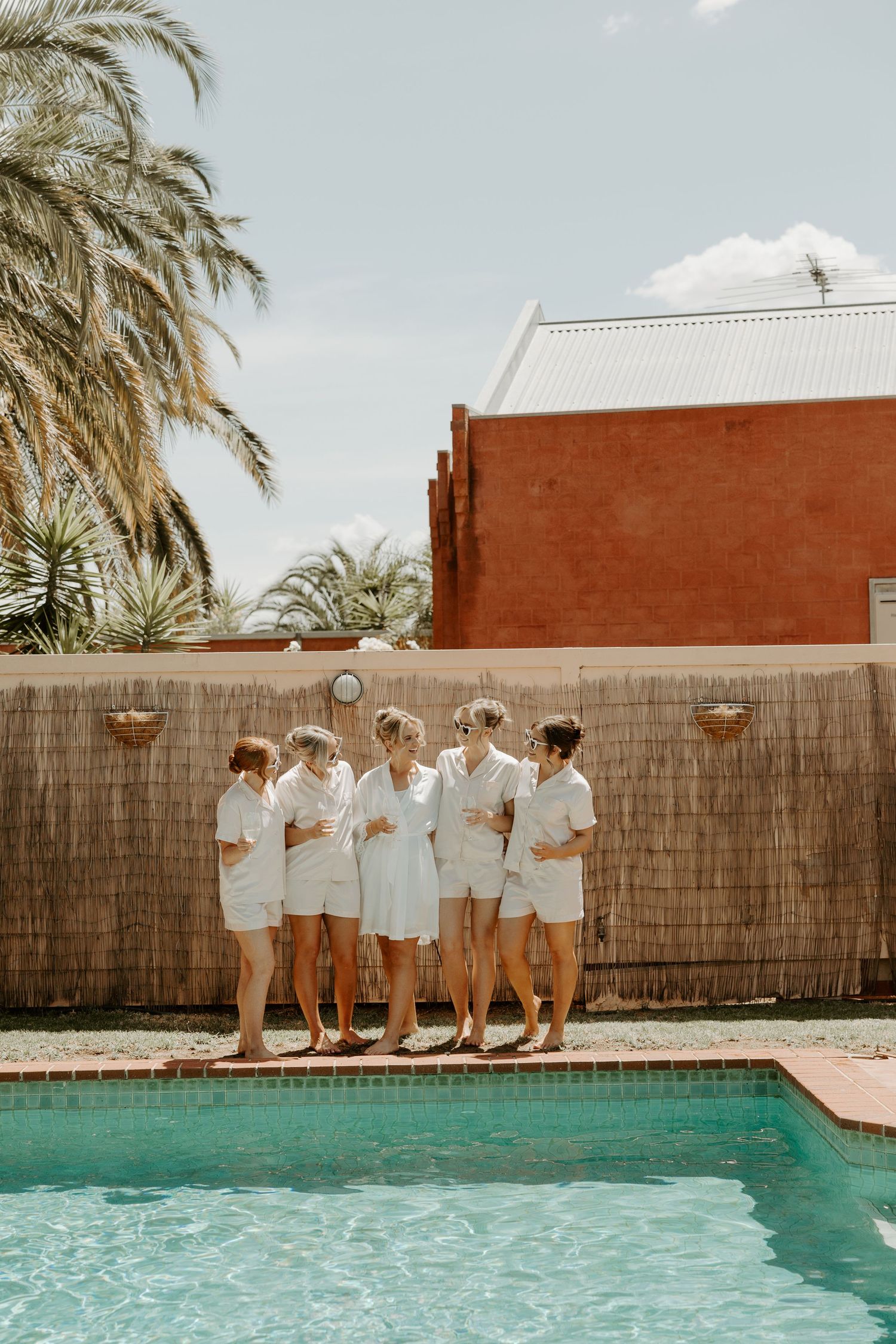 A group in white outfits stands by a pool with palm trees and a terra cotta building in a sunny resort setting.