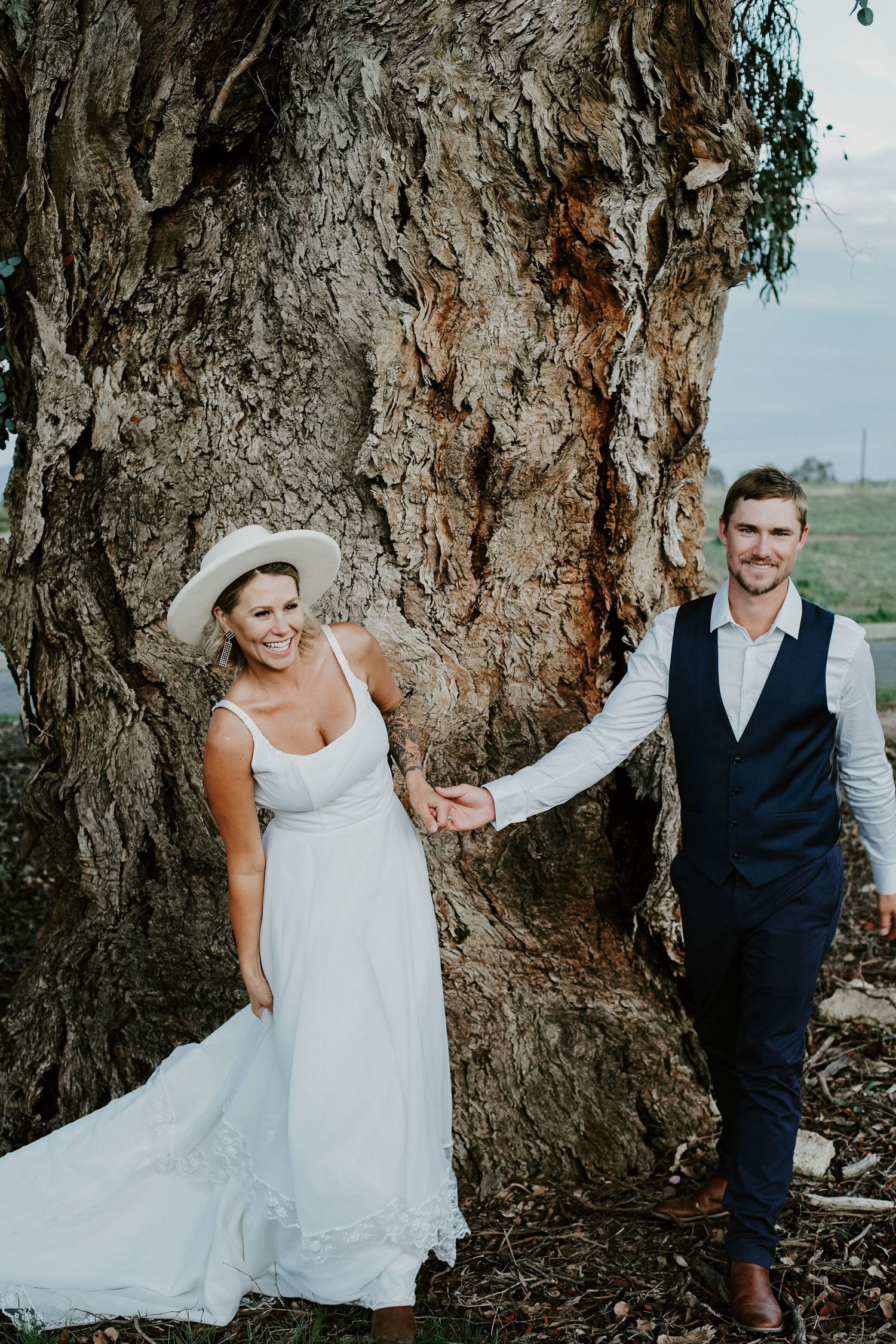 Wedding couple poses by large tree trunk in outdoor setting.