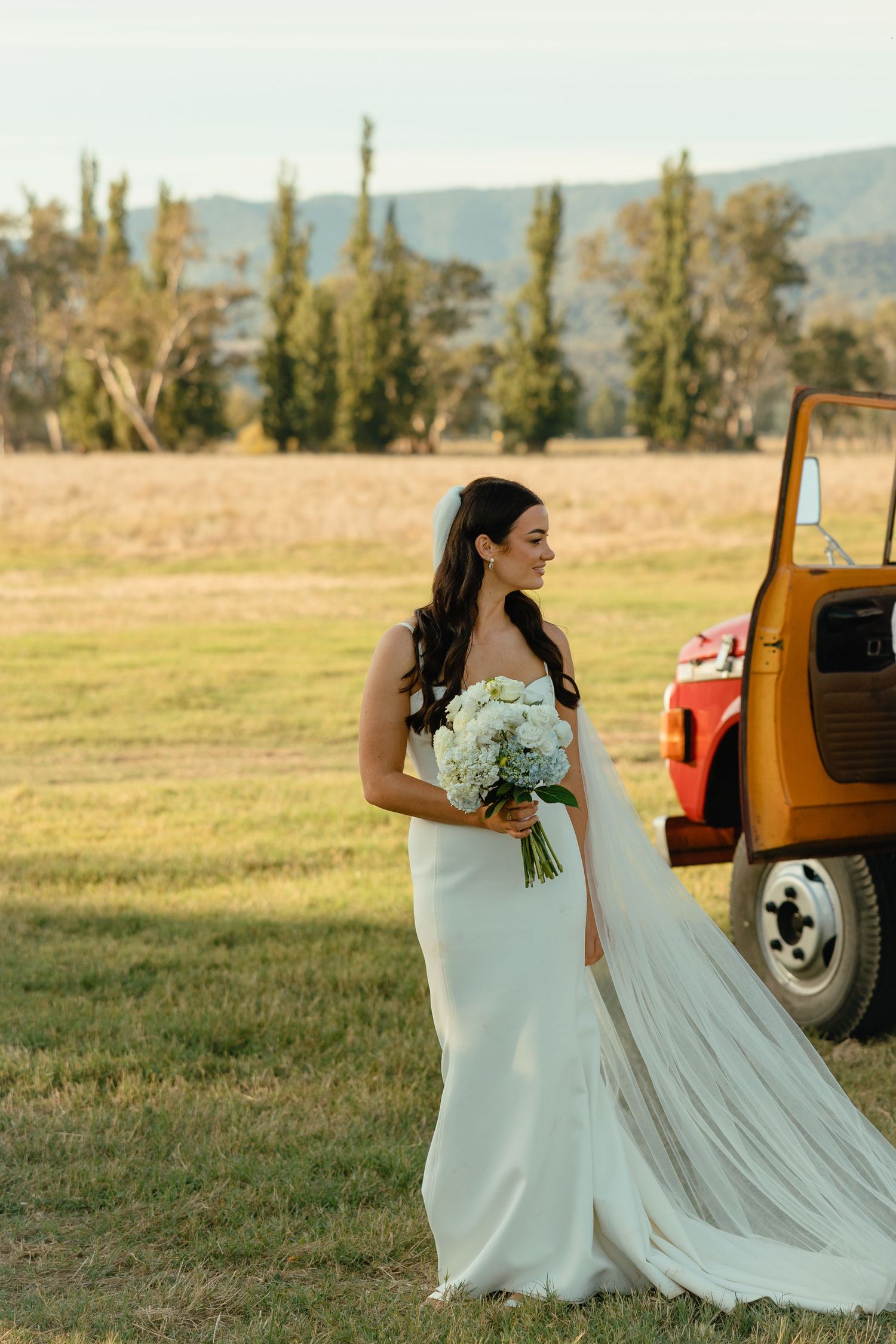 A bride in a flowing white dress holds a bouquet next to a vintage orange truck in a rural field at sunset.