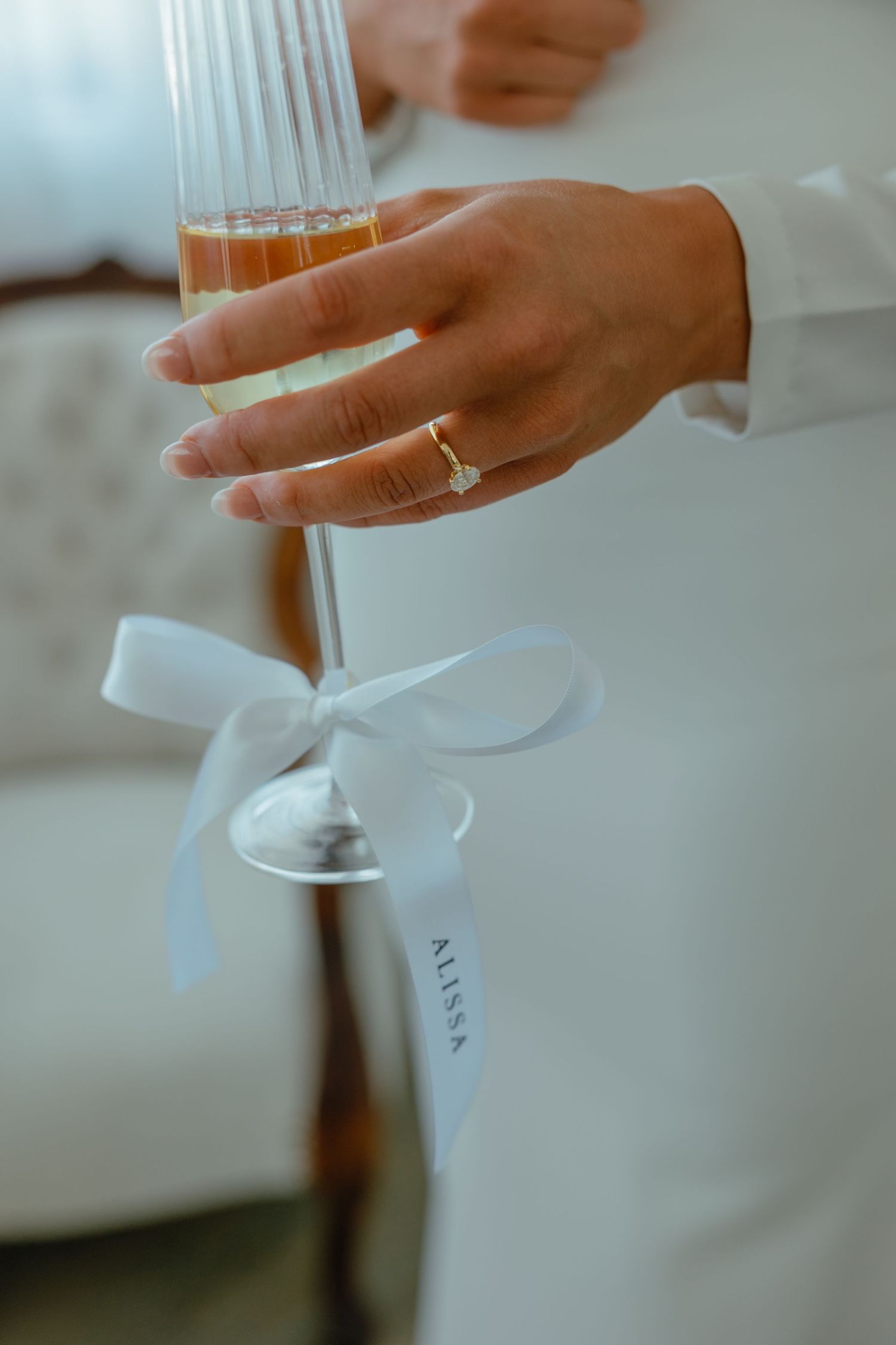 Hand wearing engagement ring holding a champagne glass with white ribbon detail.