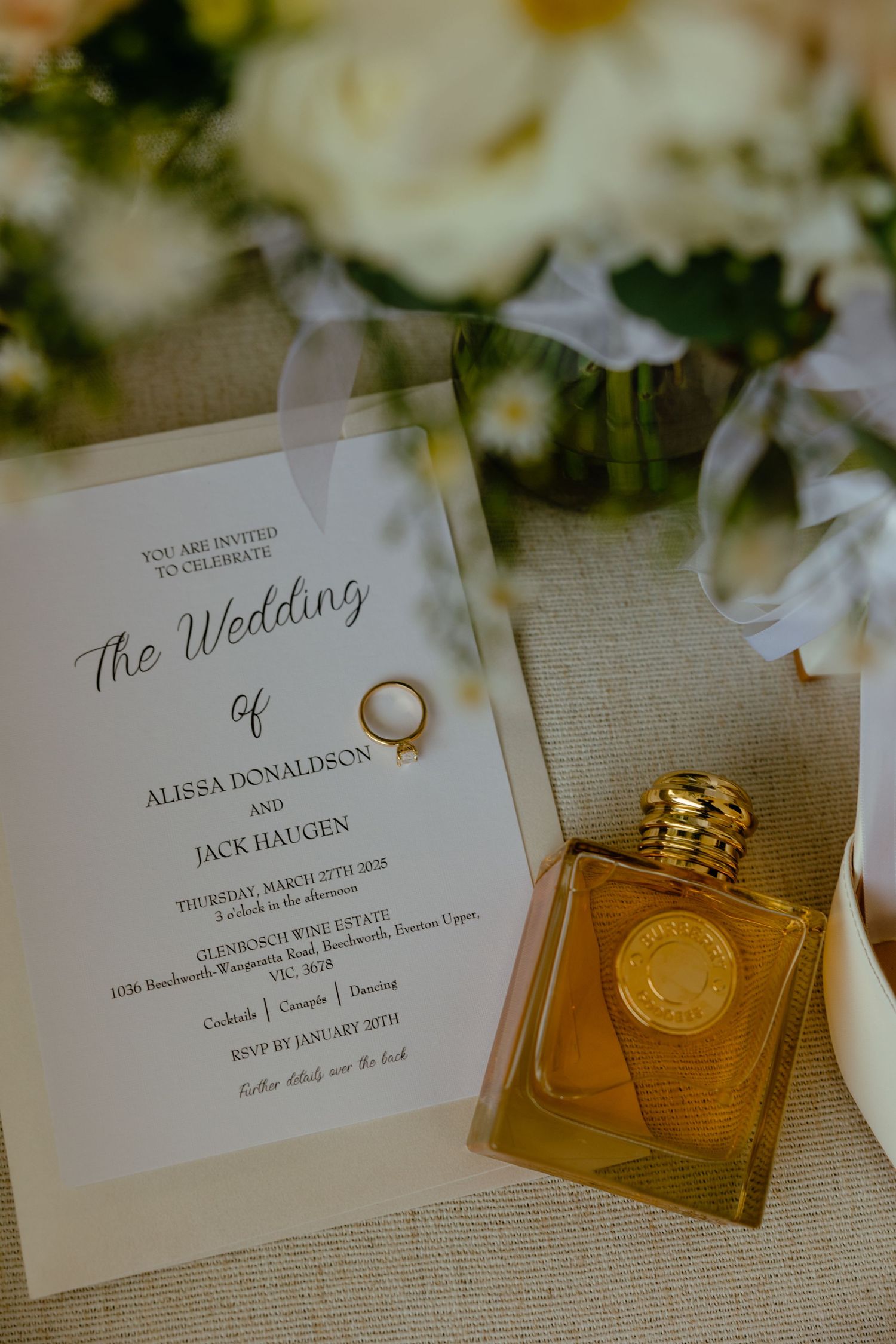 Wedding invitation and perfume bottle arranged with wedding ring capturing golden light.