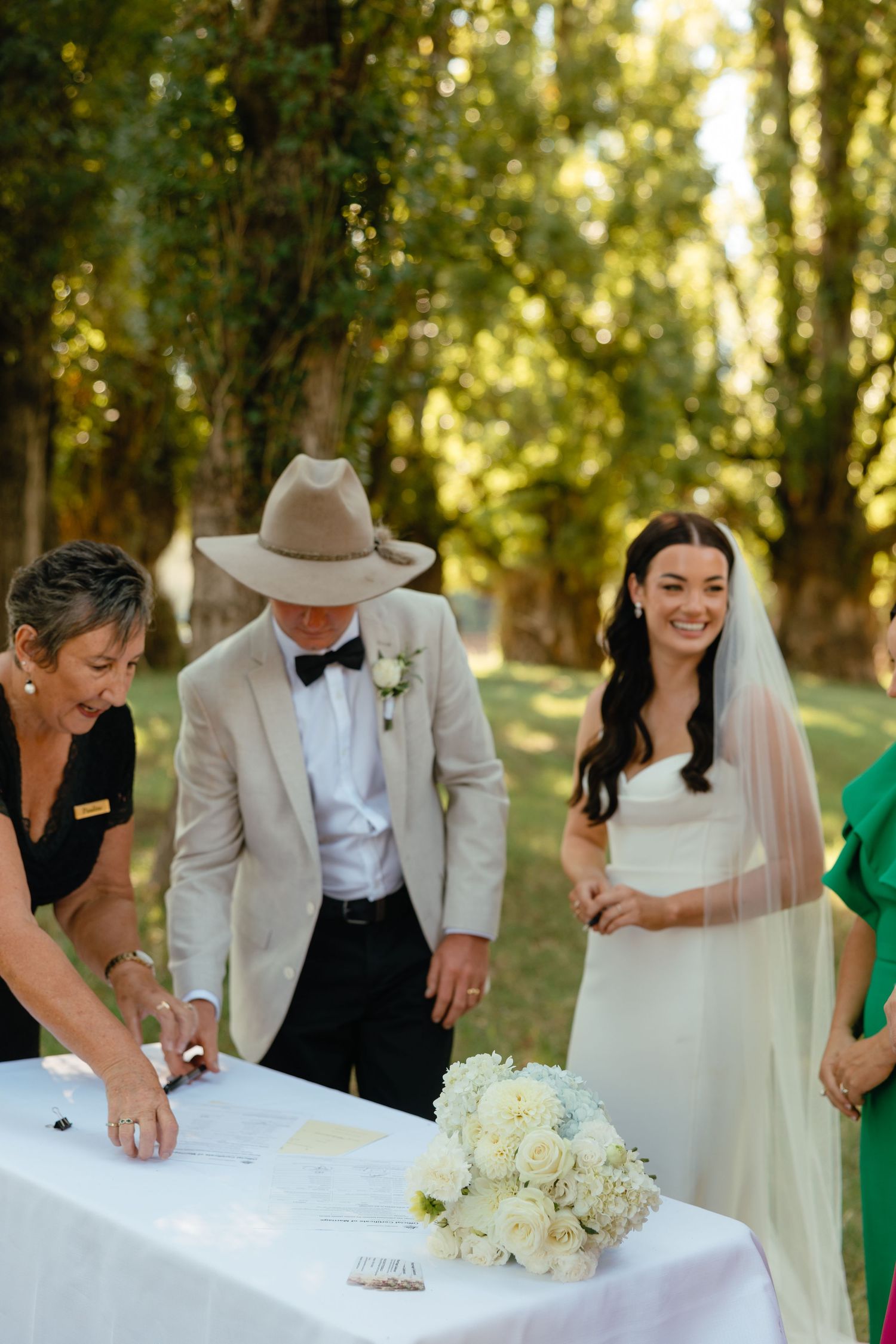 A group stands outdoors at a signing table during a wedding ceremony in a sunlit wooded setting.