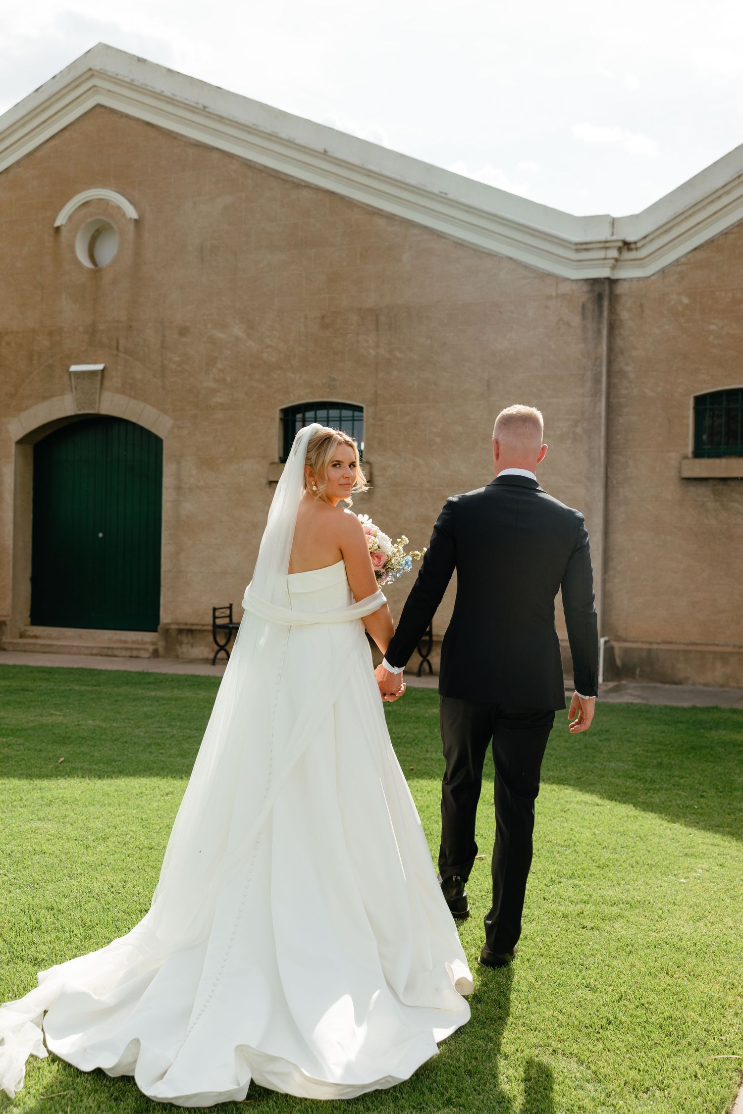 Bride in white gown and groom in black suit walking together on grass path by stone building.