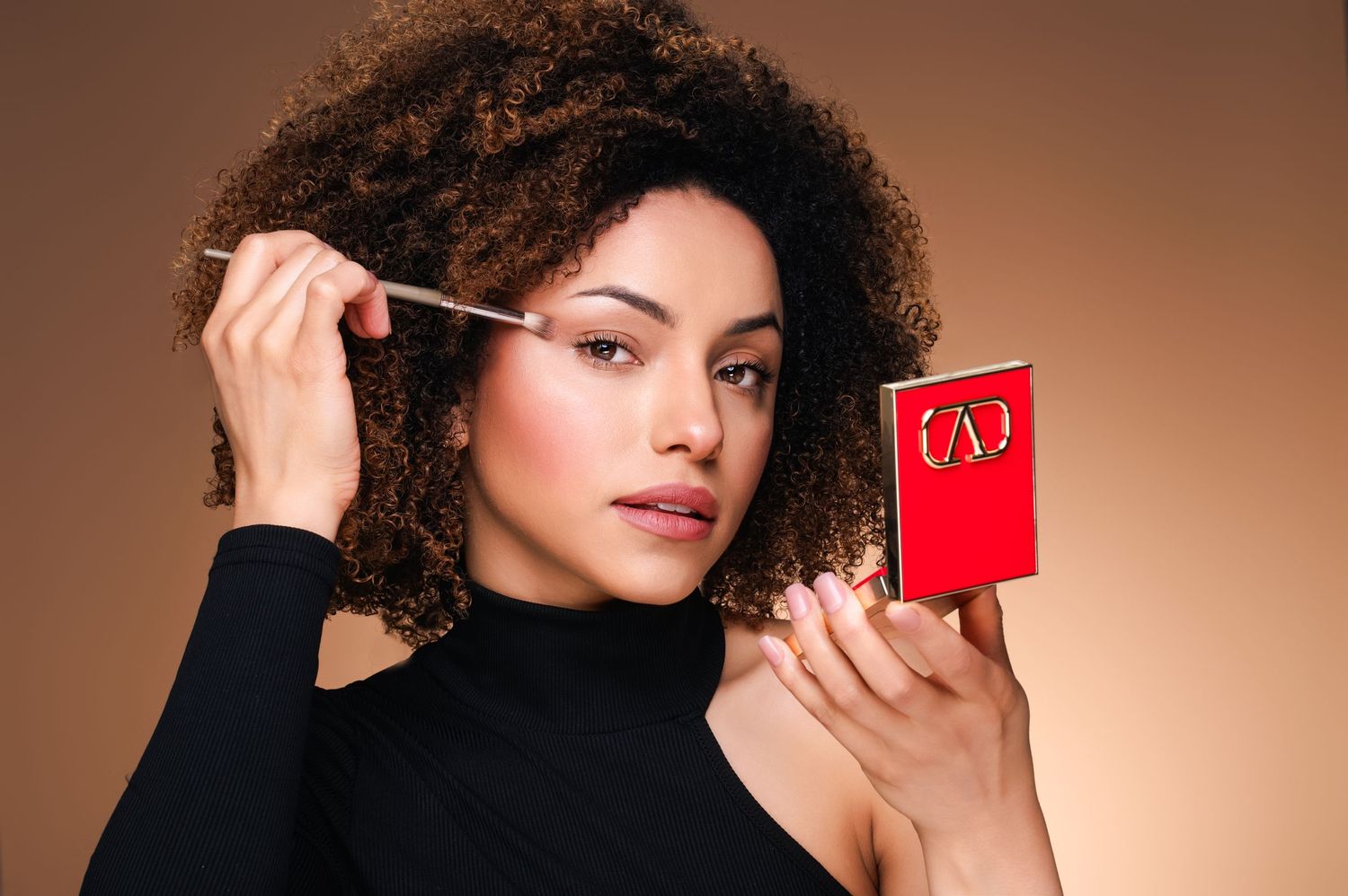A beauty shot showing someone applying mascara while holding a red compact mirror against a brown background.
