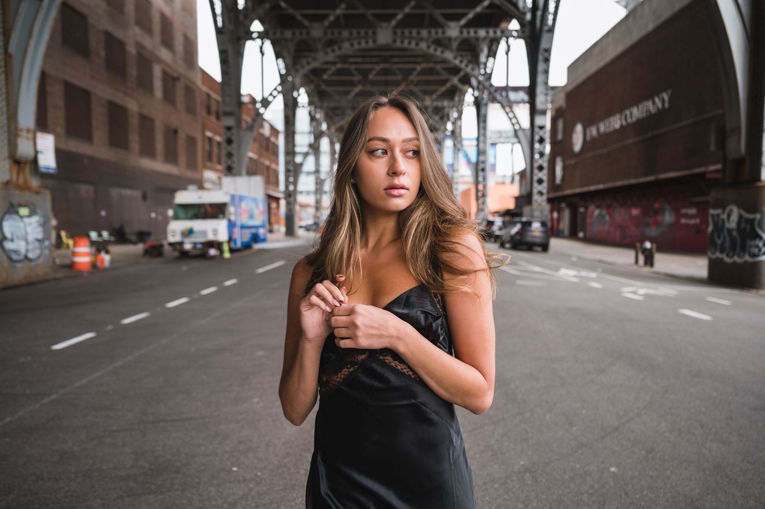 A figure stands beneath elevated train tracks on a city street with industrial architecture and buildings in the background.