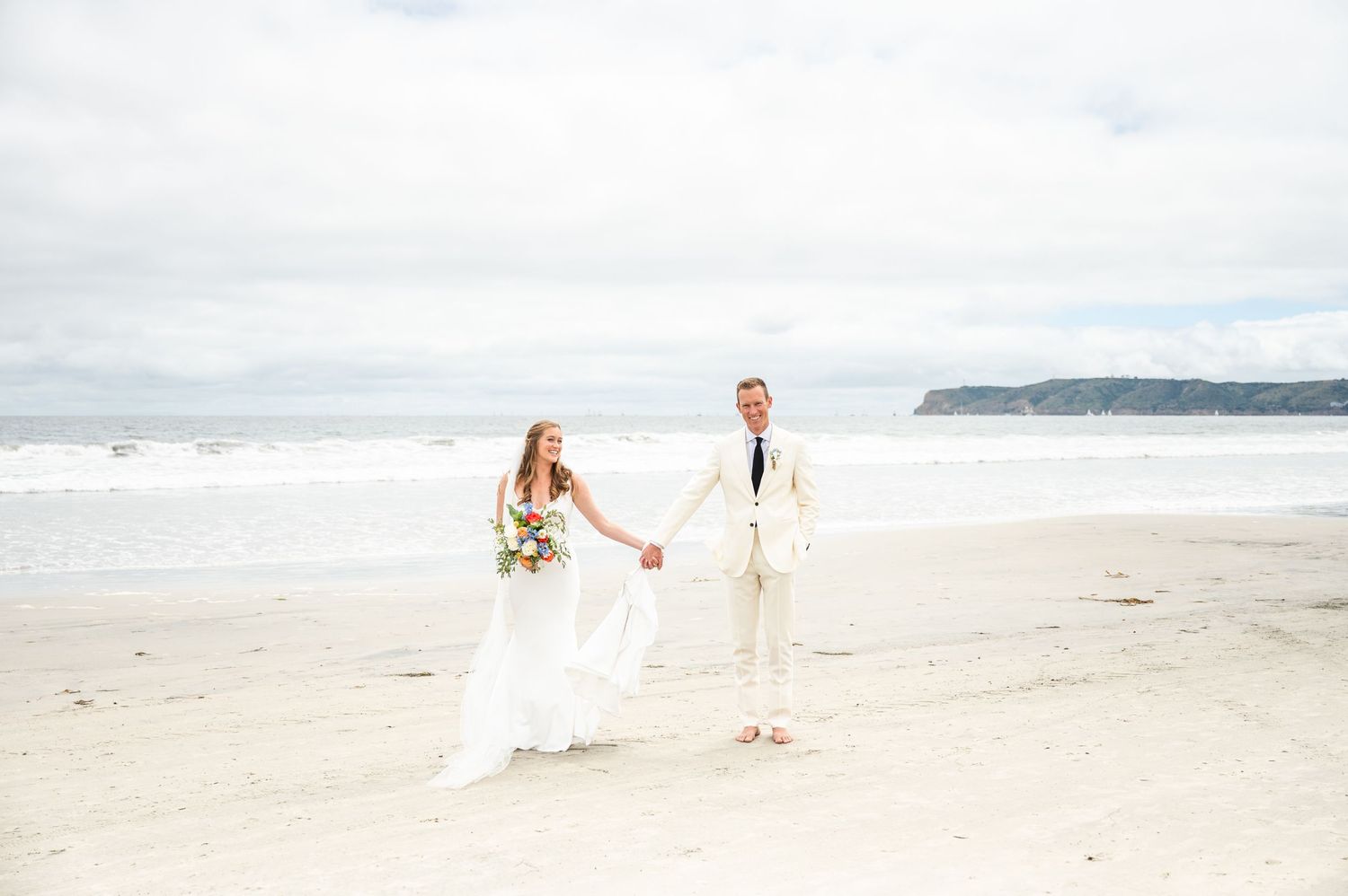 Bride and groom holding hands, walking through sand on an overcast Coronado day at the beach.