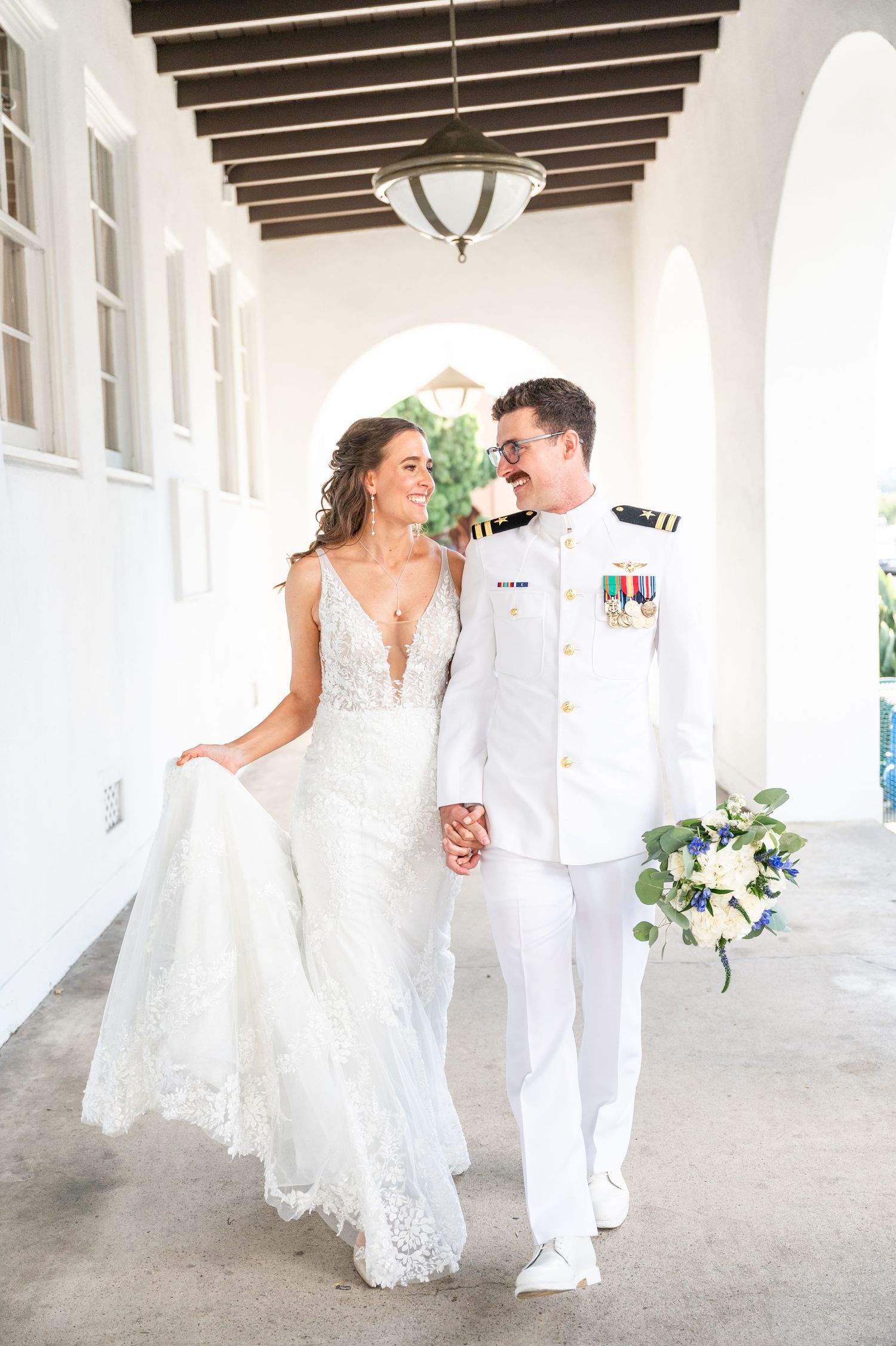 Bride in white and groom in navy pilot uniform walking side by side down a white hallway at Liberty Station Point Loma after their first loo
