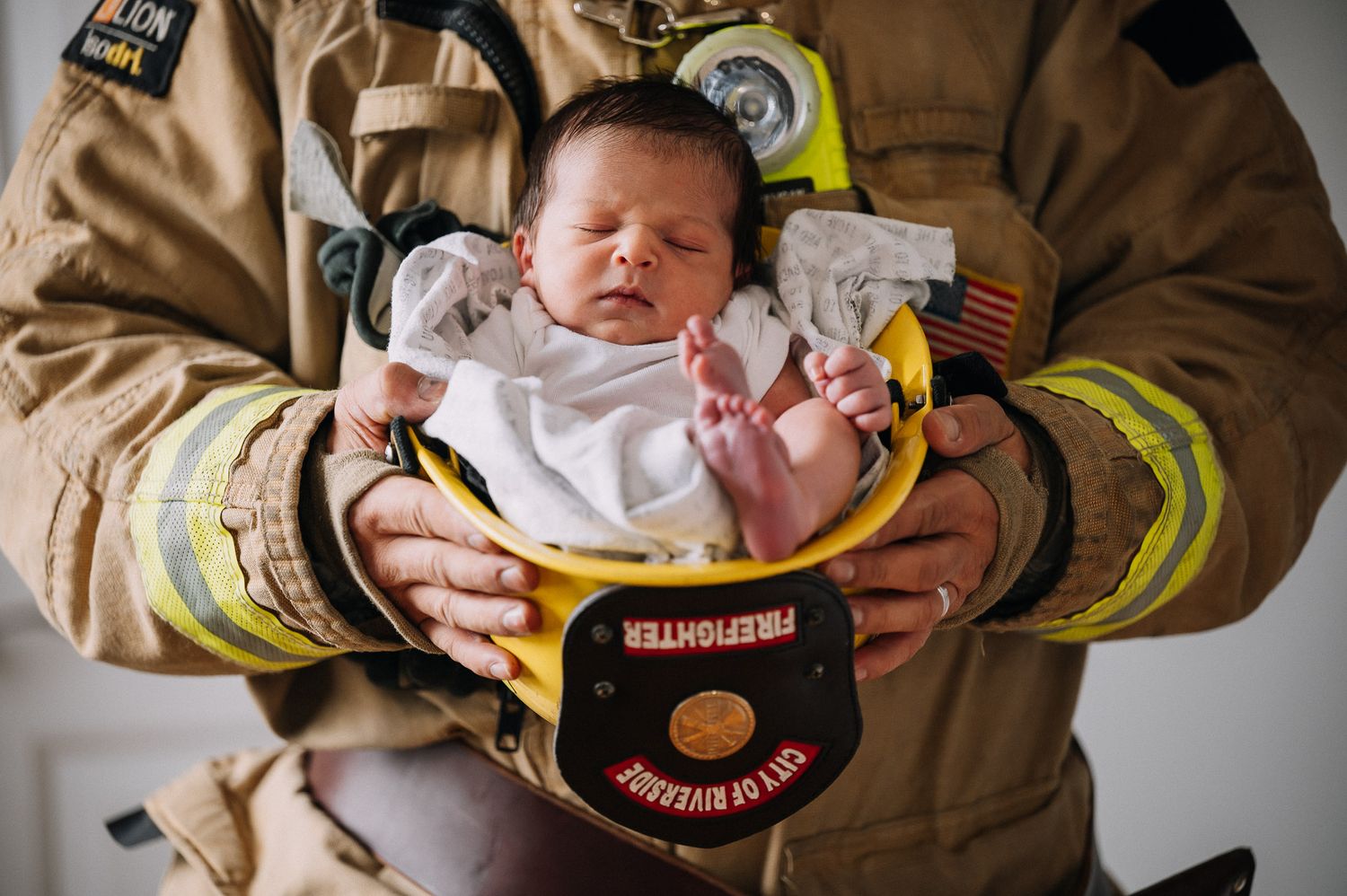 A newborn baby cradled in the arms of a firefighter wearing protective gear and department patches.
