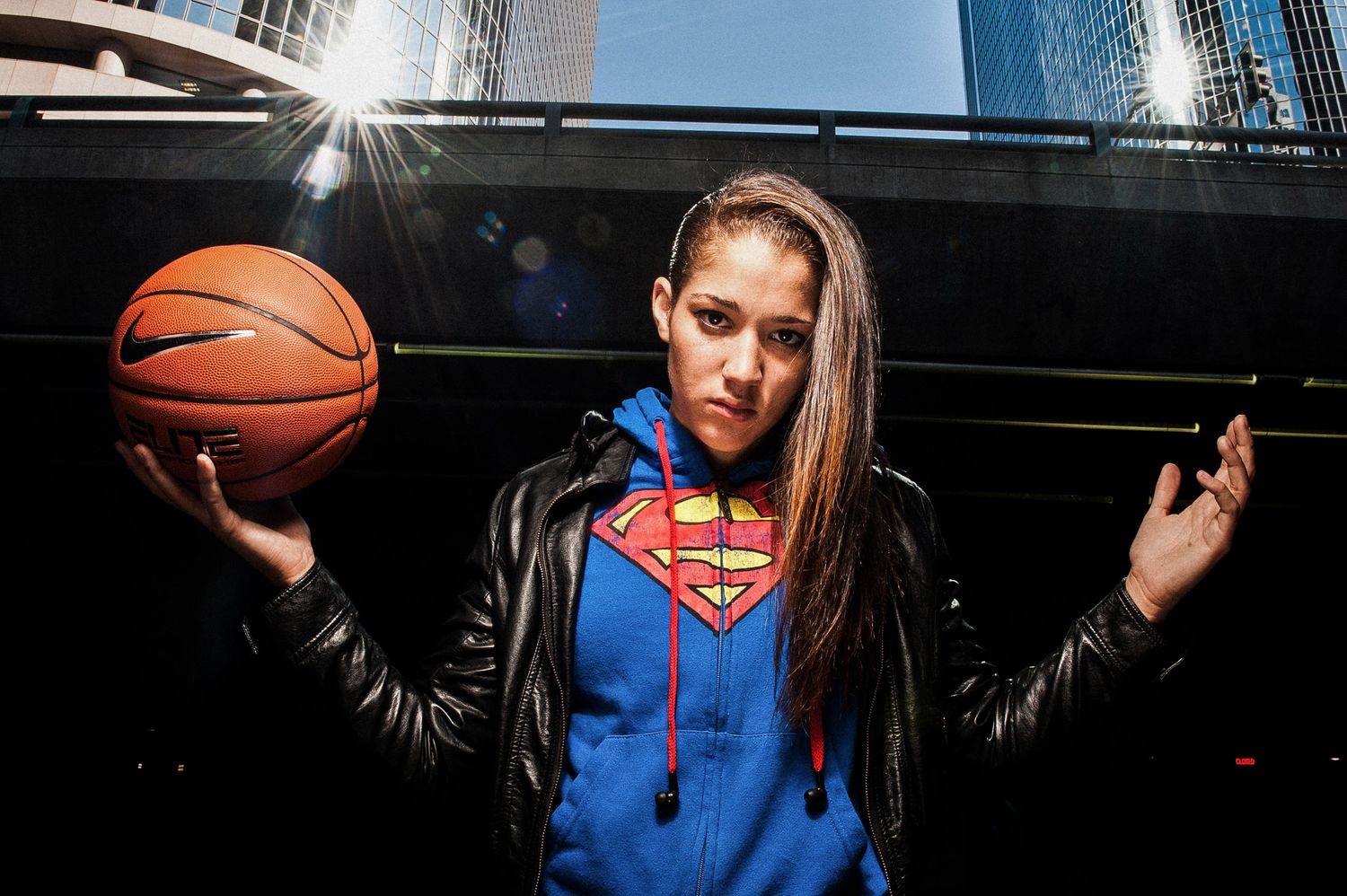 A person in a Superman hoodie holds a basketball while posing against an urban backdrop with skyscrapers.