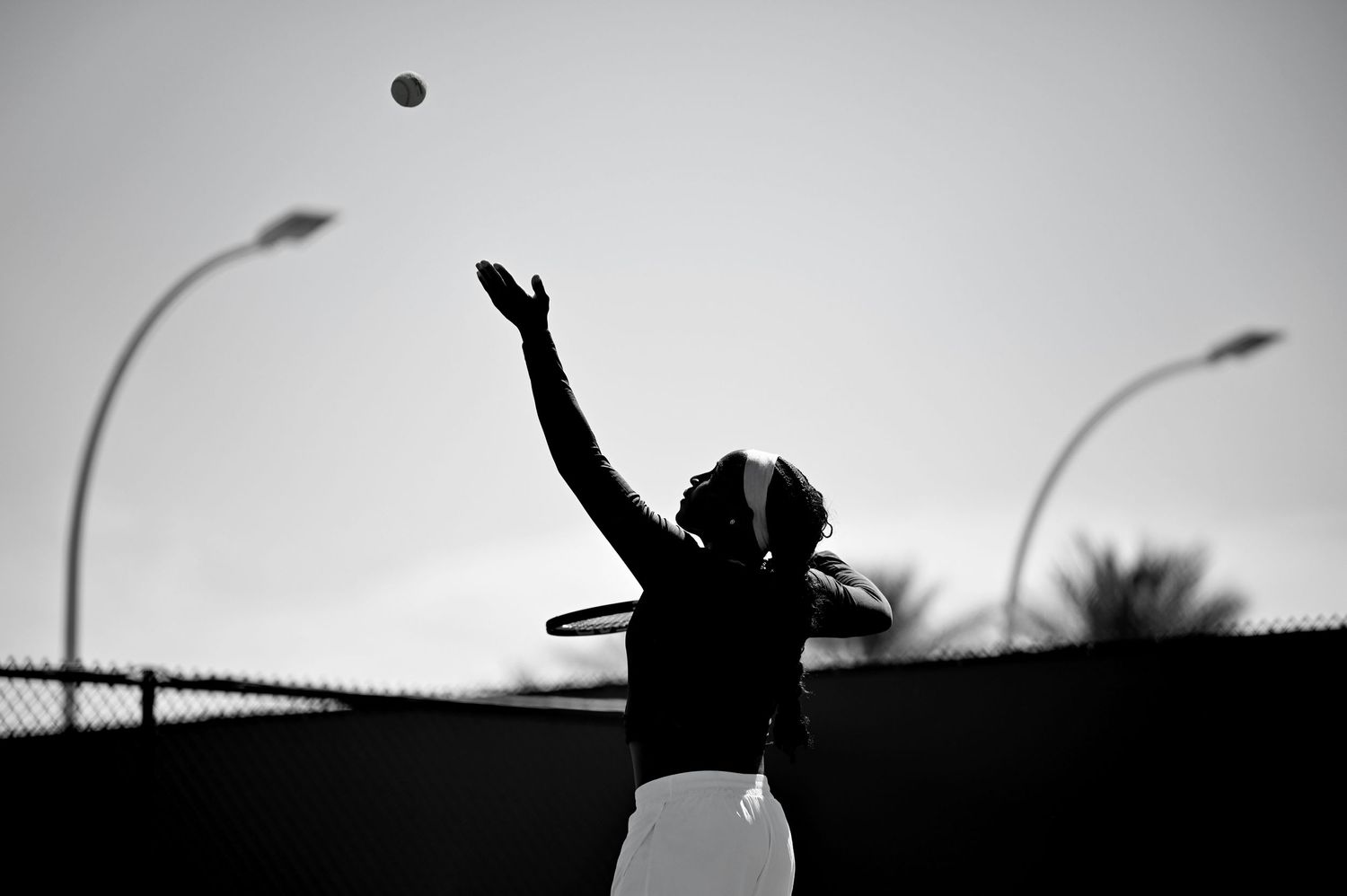 Silhouette of tennis player reaching up to serve ball against dark sky with street lamps in background.