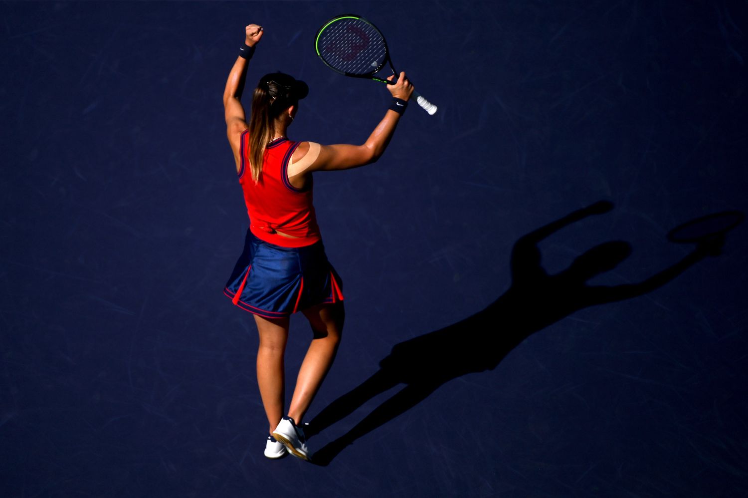 Tennis player in red shirt and blue skirt serves while casting a dramatic shadow on the court.