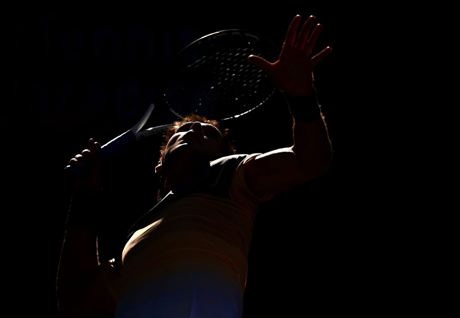 Tennis player silhouette serving ball against dark background with dramatic rim lighting.