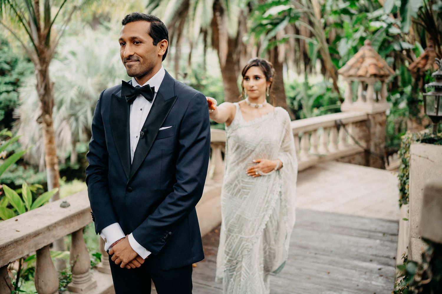 A couple in formal wedding attire stand on a garden pathway surrounded by tropical palm trees and greenery.