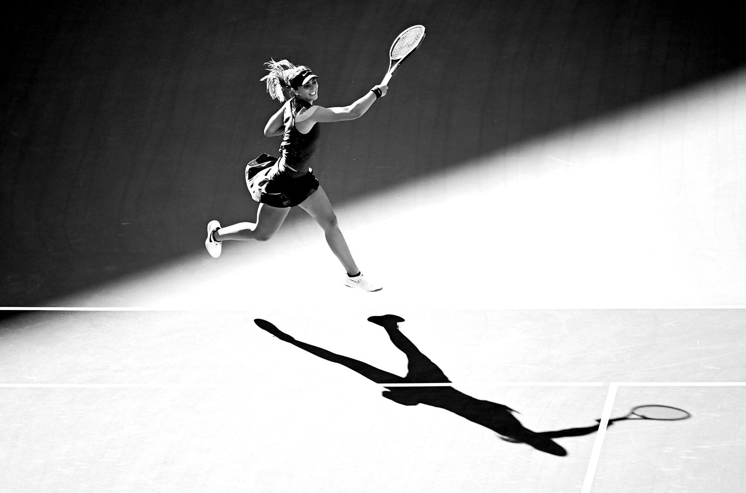 A dramatic black and white photo of a badminton player leaping to make a shot, with a distinct shadow below.