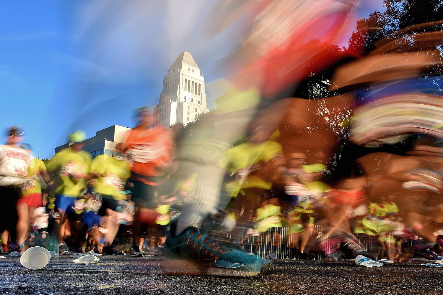 Motion blur of marathon runners racing past a white building on a sunny day.