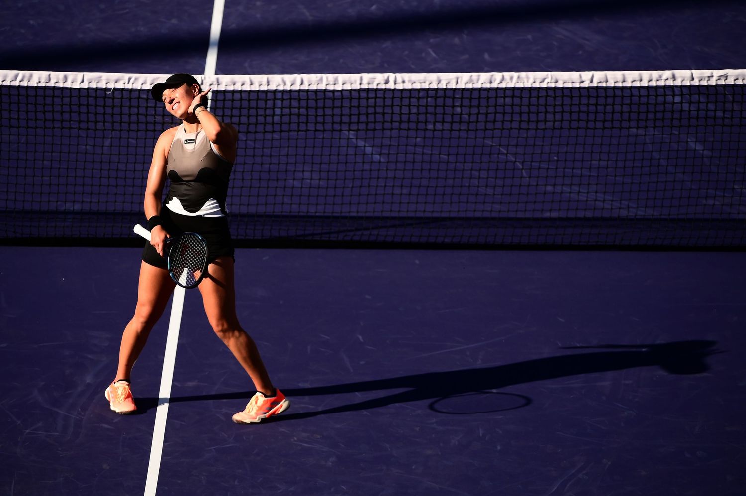 Tennis player casts long shadow on purple court while walking across white line during sunny day.
