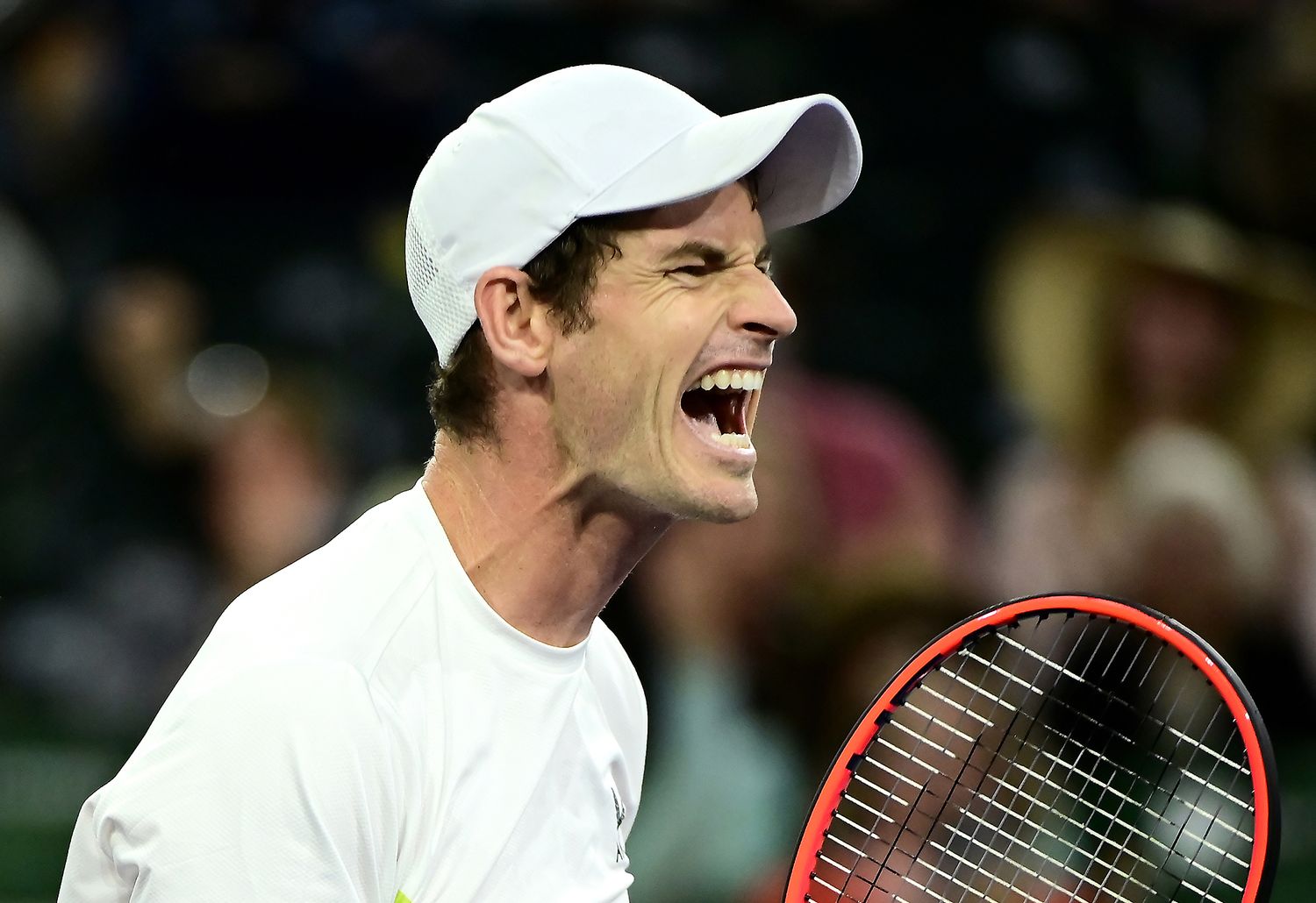 Tennis player wearing white clothing and cap celebrates emotionally with mouth open during a match.