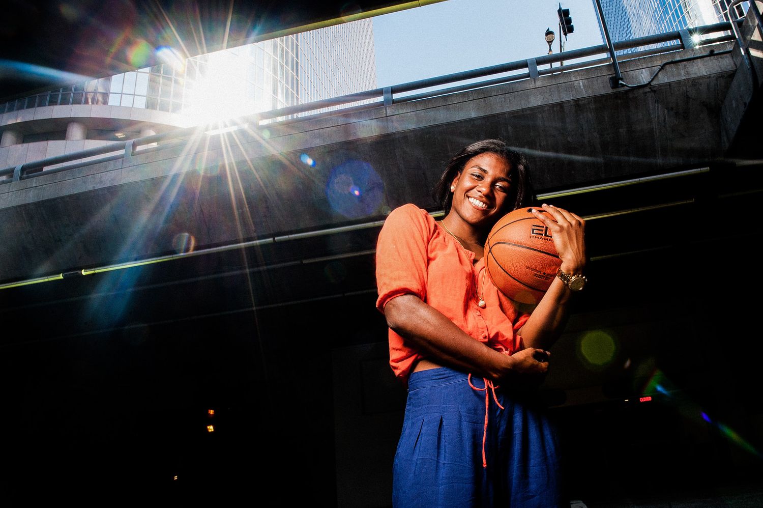 Basketball player in orange shirt holds ball while standing on outdoor court with dramatic sunburst lighting overhead.